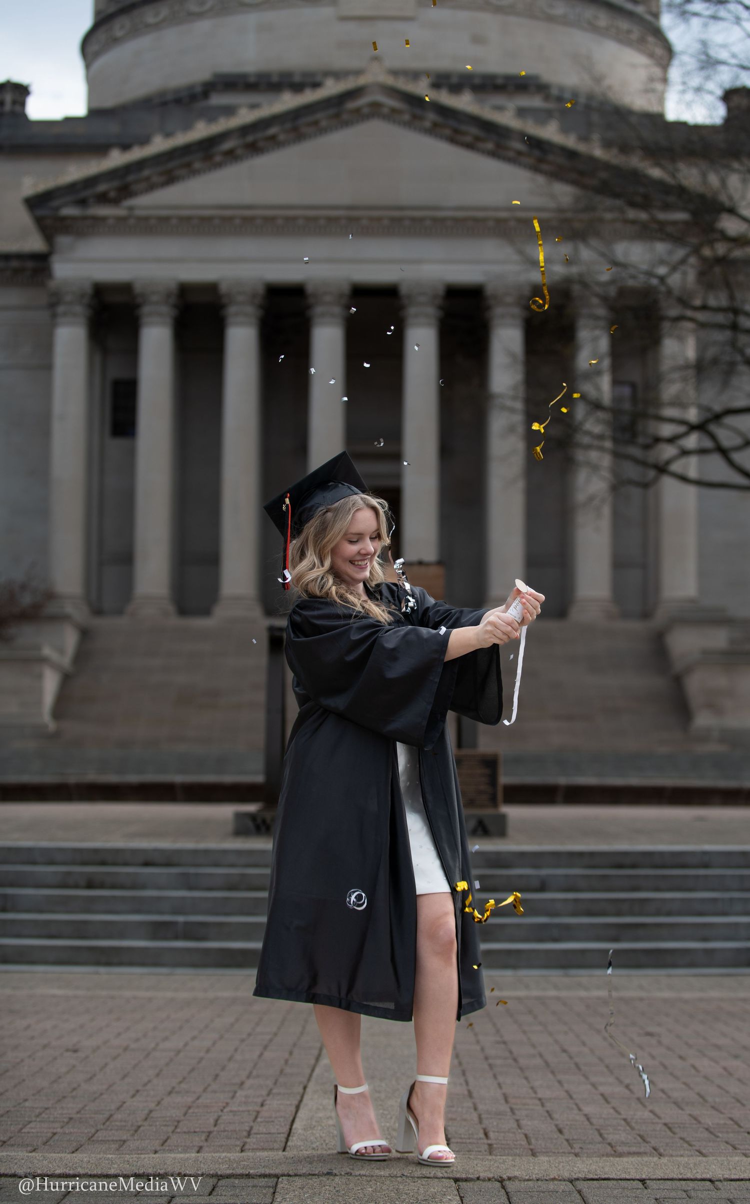 A student in graduation attire celebrates by tossing glitter and confetti outside a columned university building.