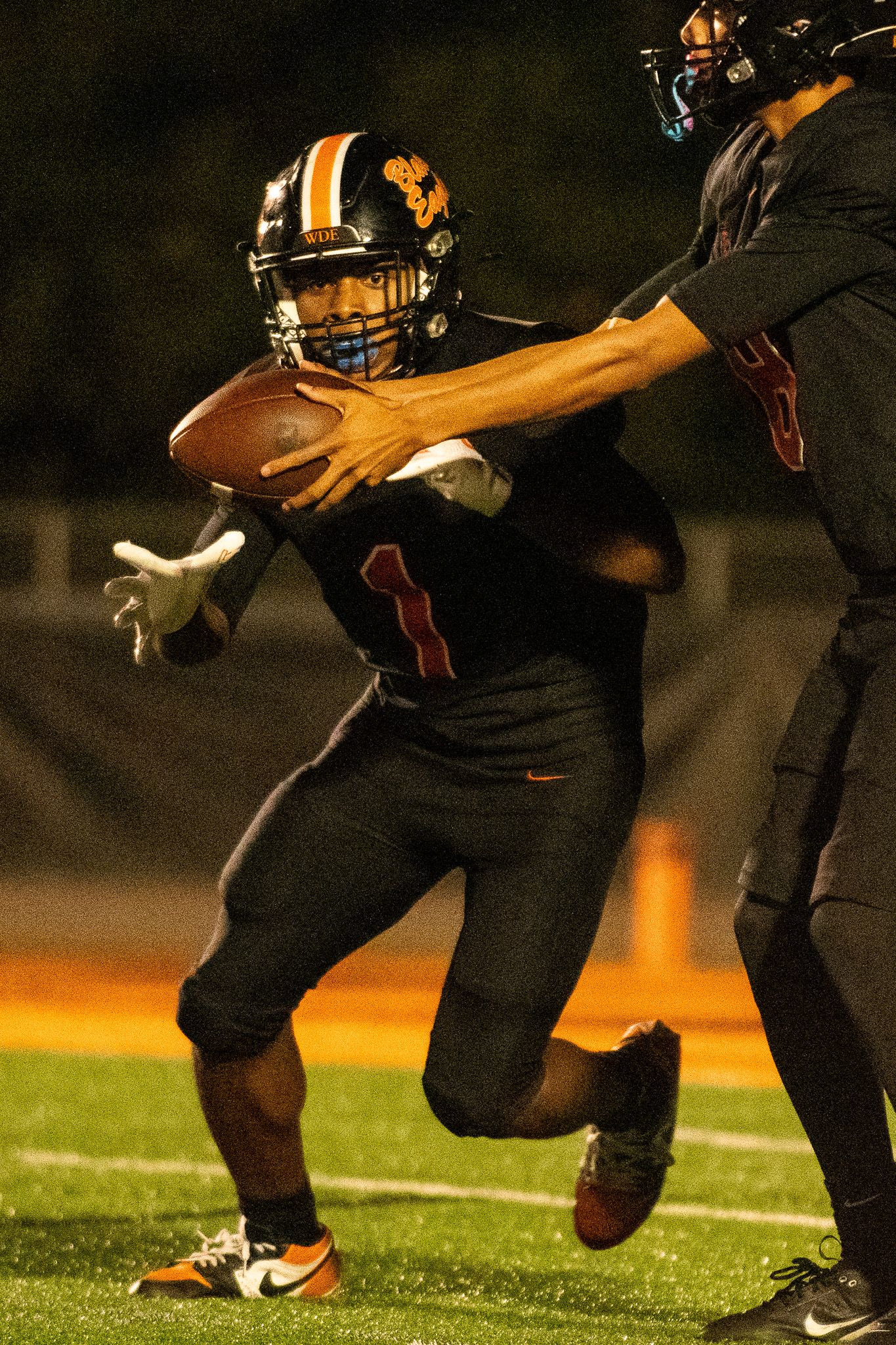 Football player in black uniform makes dynamic running motion across field during nighttime game under stadium lights.