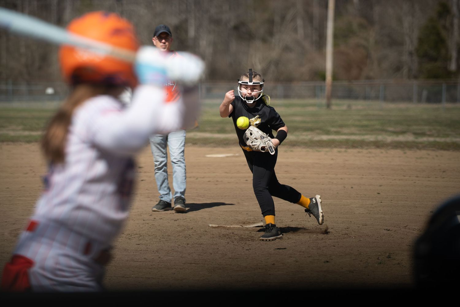 Youth softball game action with pitcher delivering pitch to batter at home plate.