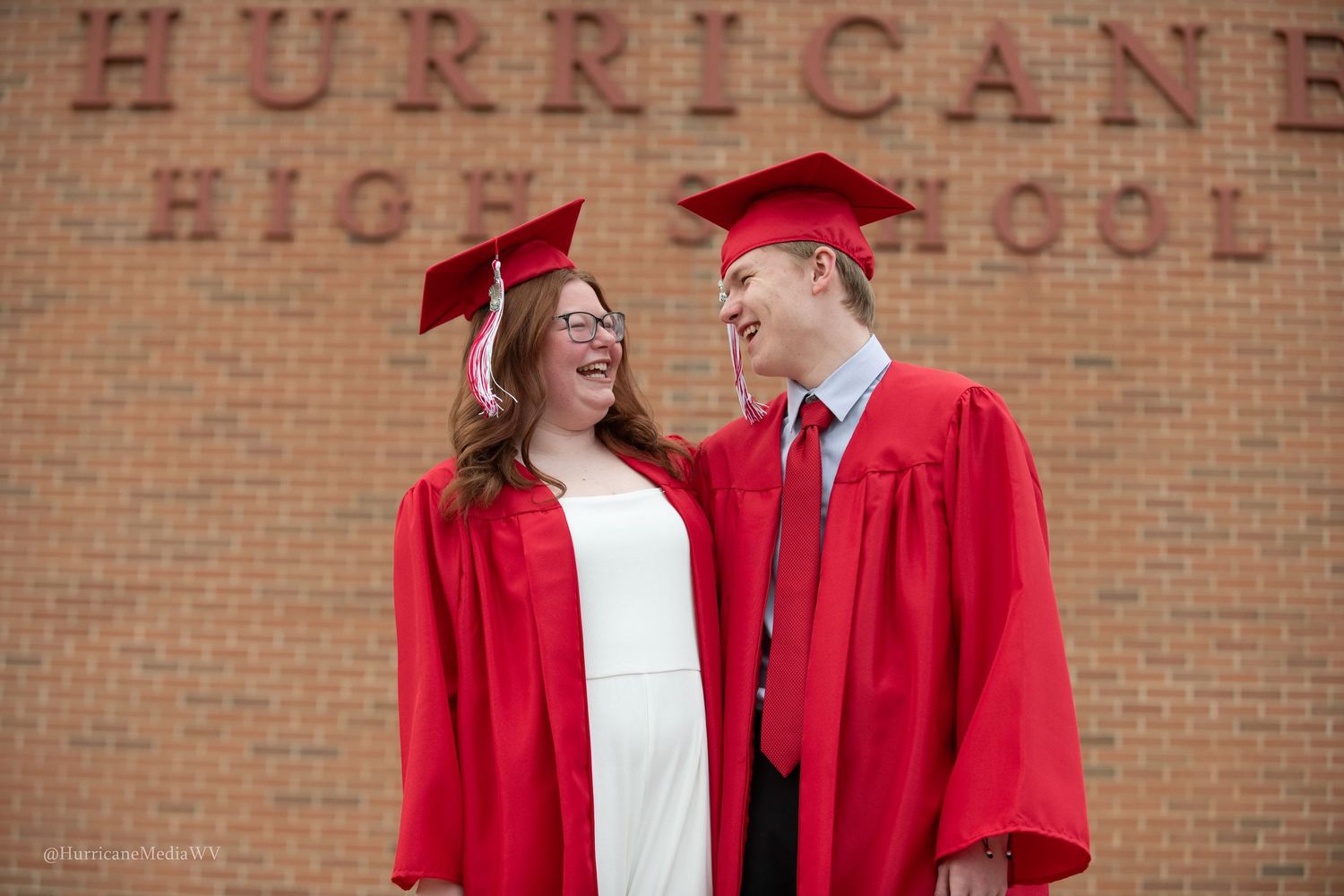 Students in red graduation gowns stand in front of a brick building with Hurricane High sign.
