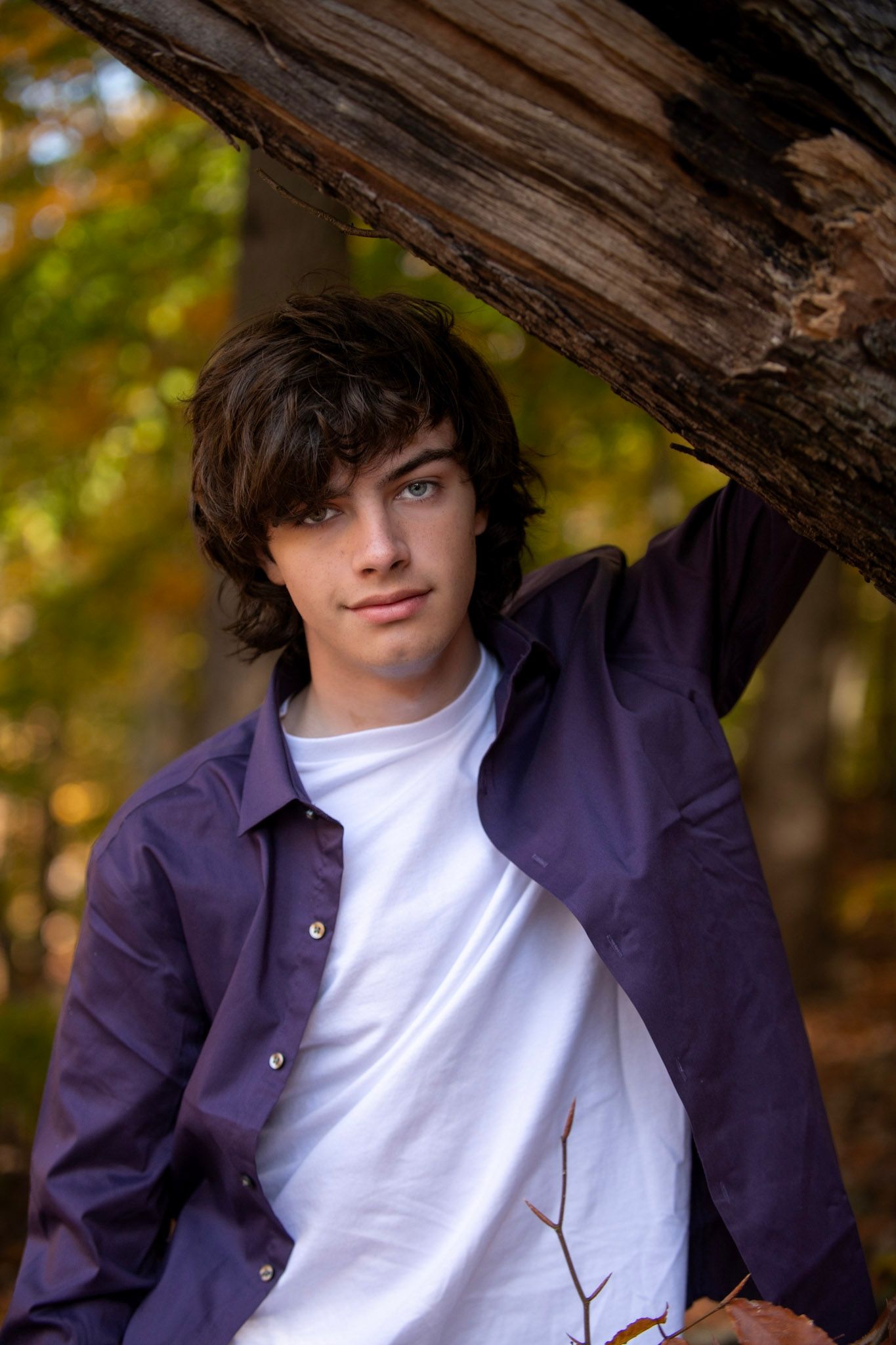 Person in navy jacket and white shirt poses casually against tree trunk in autumn setting.