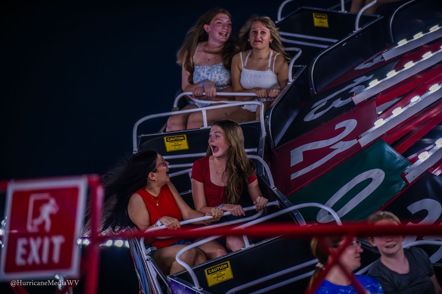 Students enjoying a thrilling ride on a roller coaster at night with red exit signs visible.