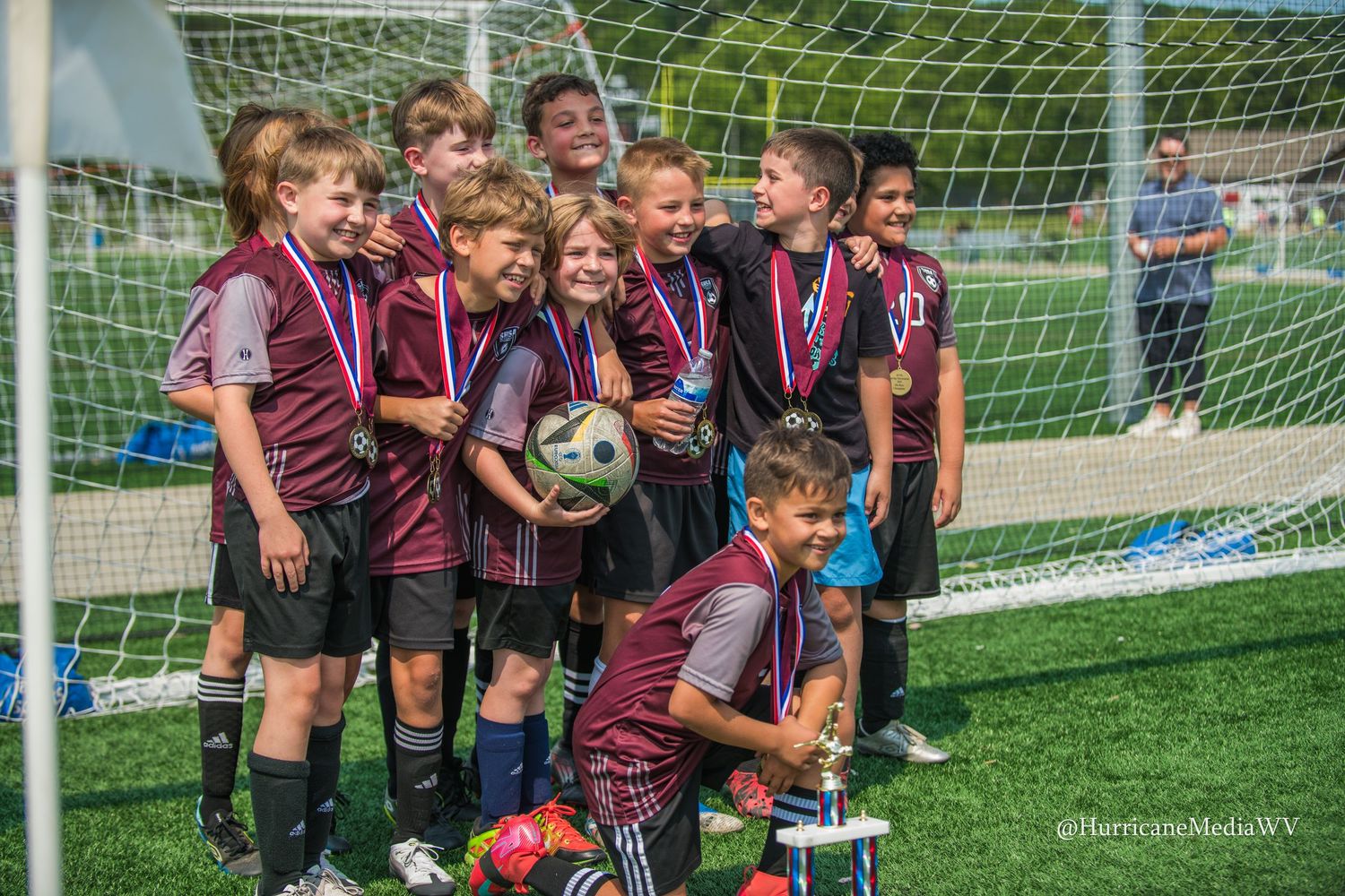 Young athletes in maroon uniforms gathered by a soccer goal celebrating together on a sunny day.