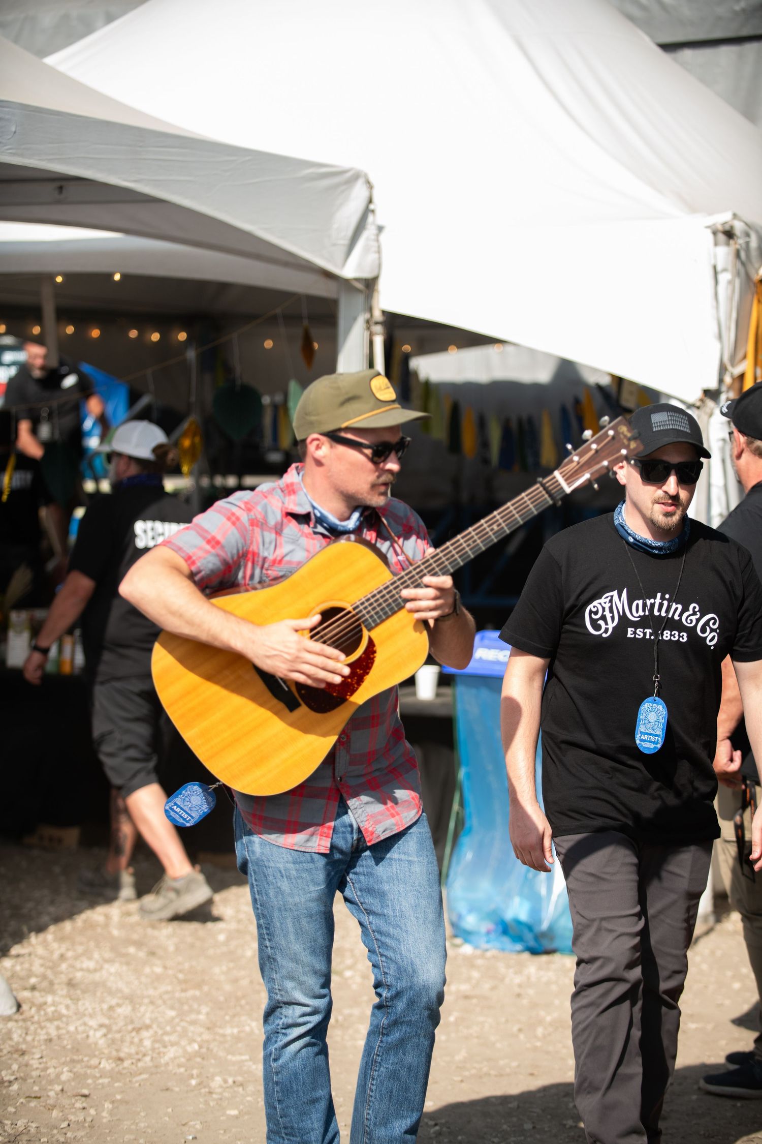Musicians perform with acoustic guitars under a white tent at an outdoor festival event.
