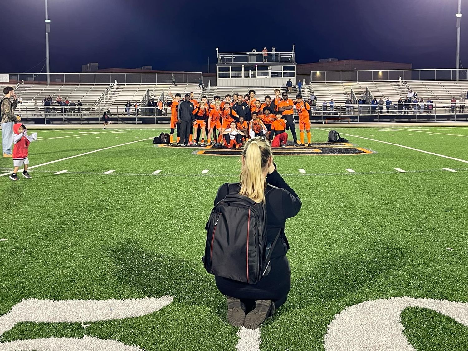 A photographer on a football field captures a team huddle under stadium lights at night.