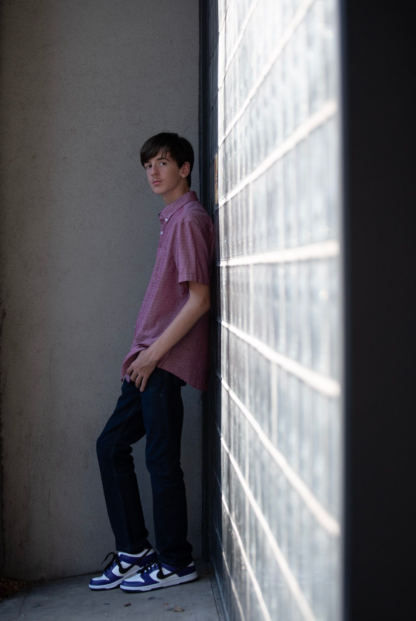 Figure in purple shirt leans against white wall with dramatic shadow patterns.