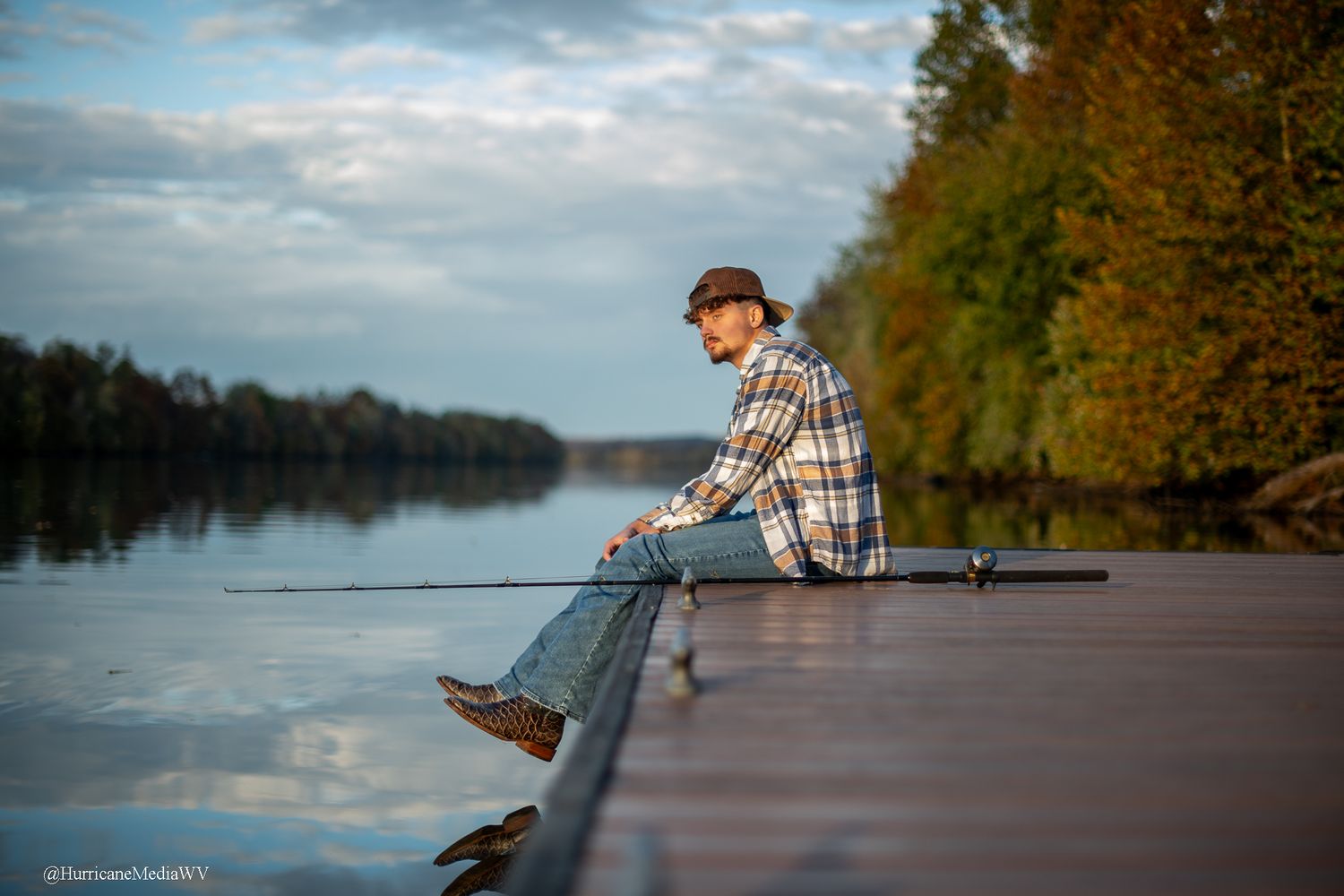 A fisherman in plaid shirt casts a line from a wooden dock on a serene lake during autumn.