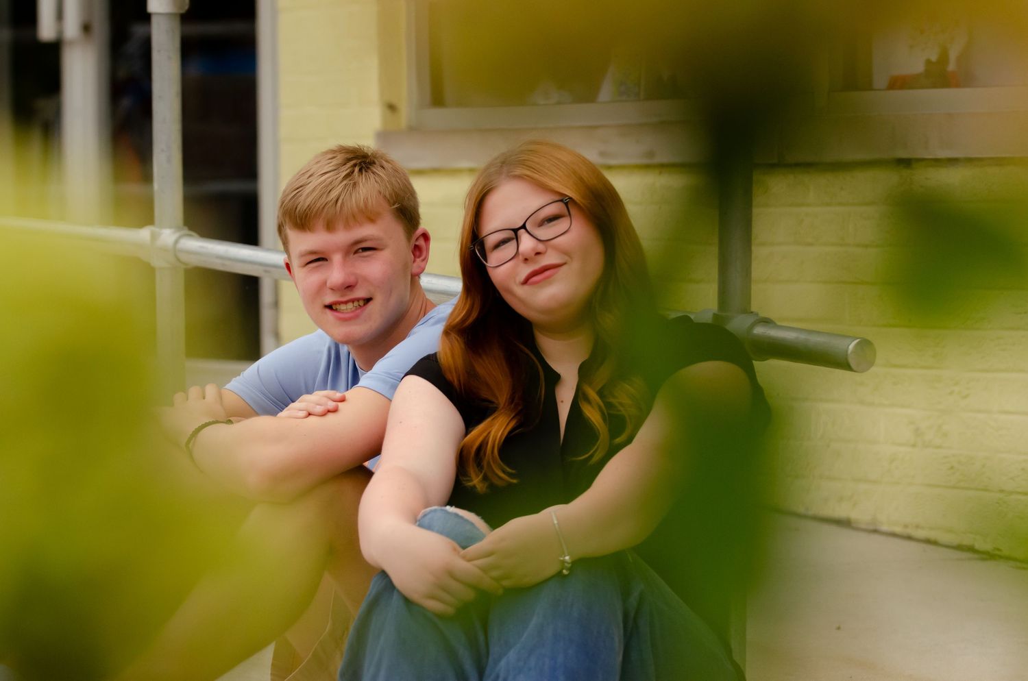 Friends share a casual moment on outdoor bench through green foliage.