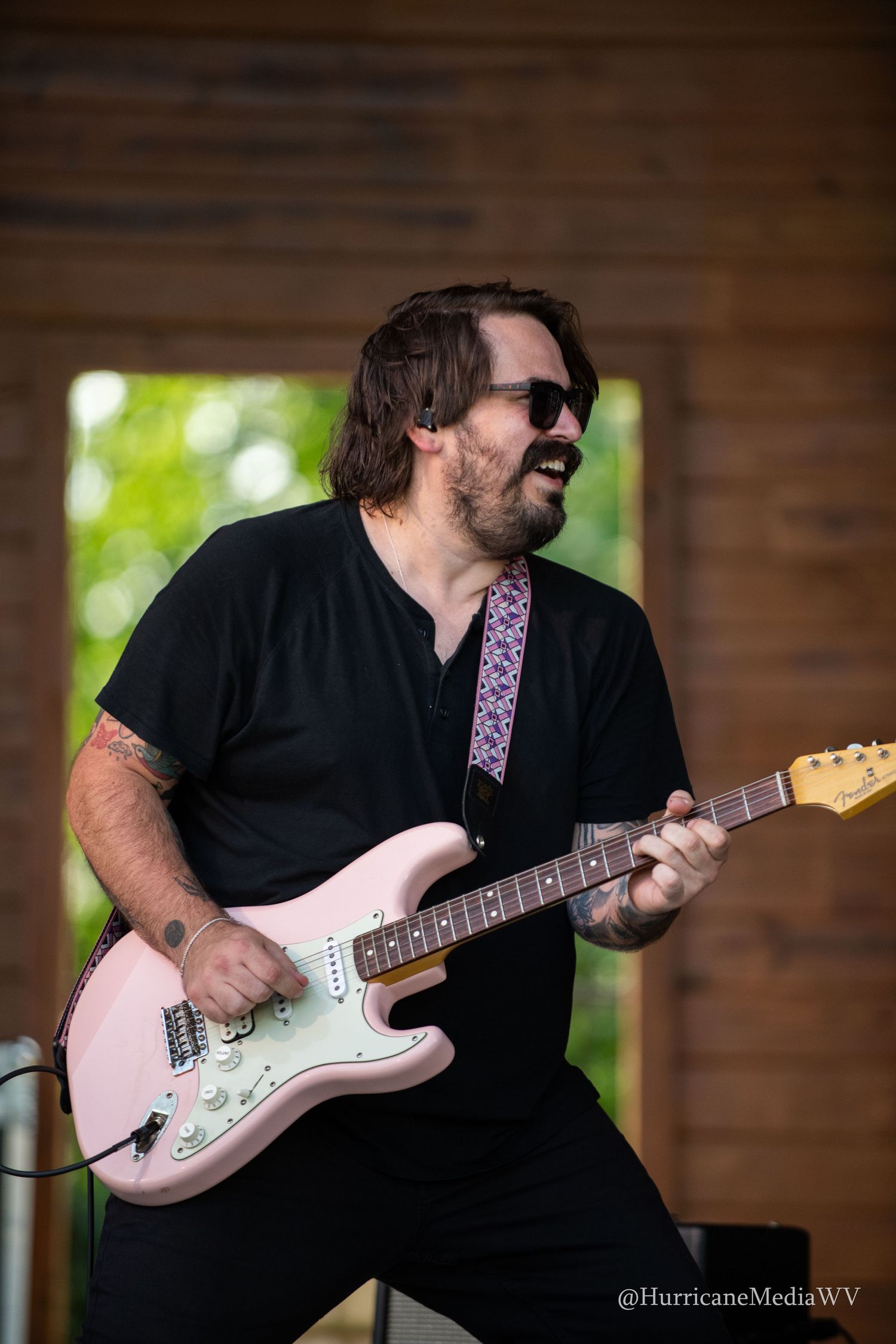 A guitarist wearing black performs on stage with a light-colored Fender Stratocaster electric guitar against a wooden backdrop.