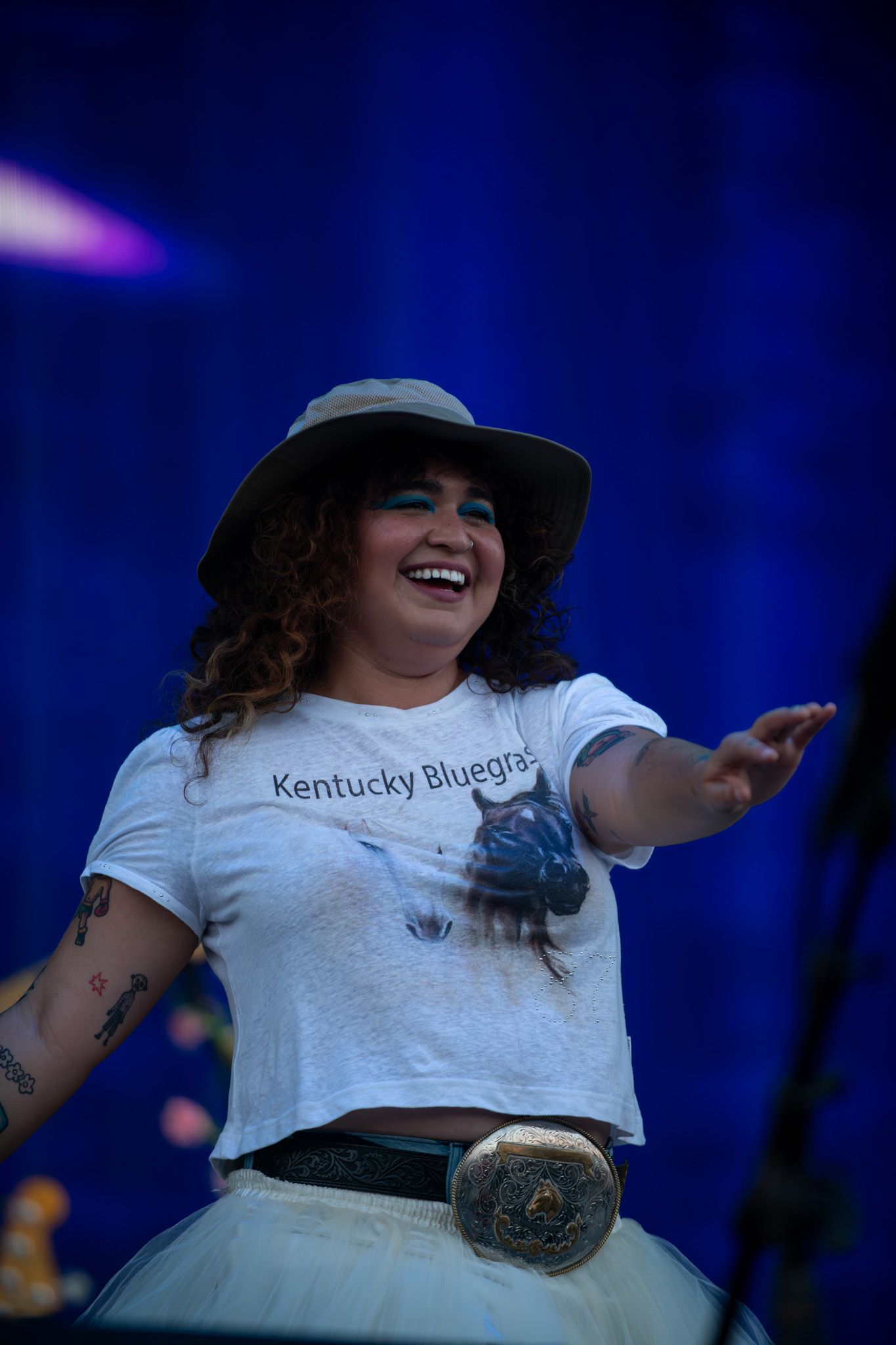 A performer in a white shirt and dark hat gestures energetically on stage under blue lighting.