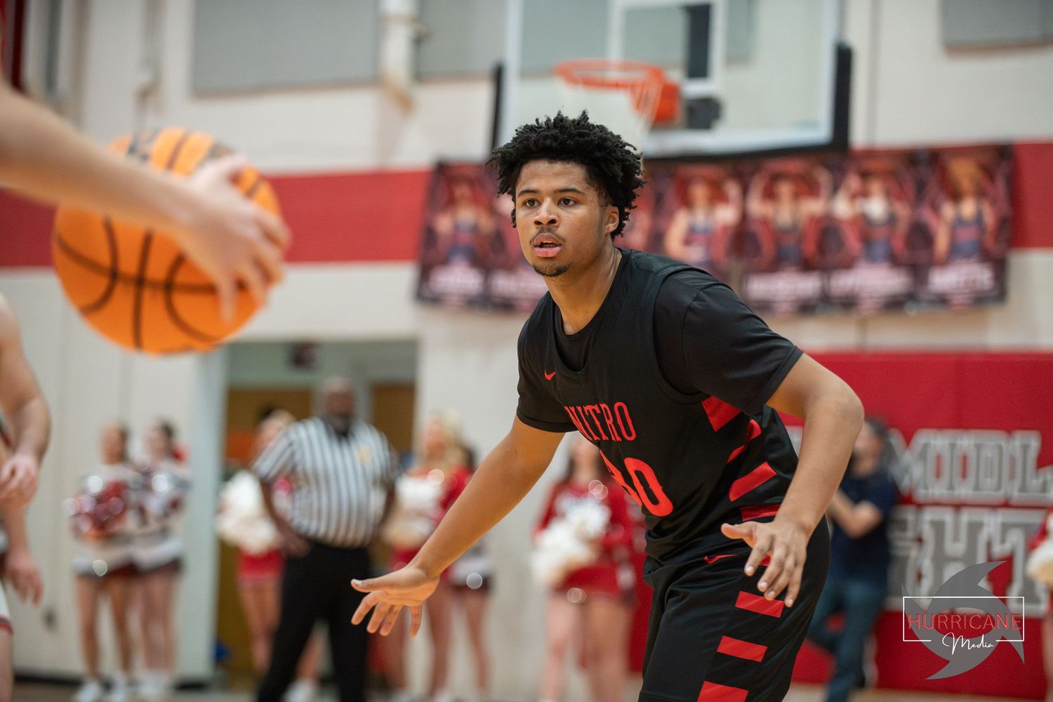 Basketball player in black jersey defends against opponent during high school game in red and white gymnasium.