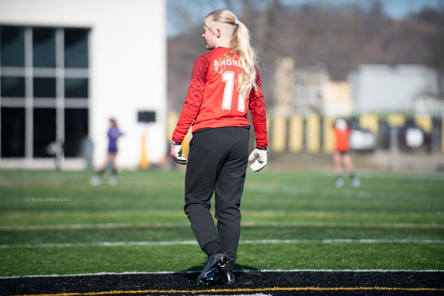 Goalkeeper in red jersey and black pants stands on soccer field looking upward during game.