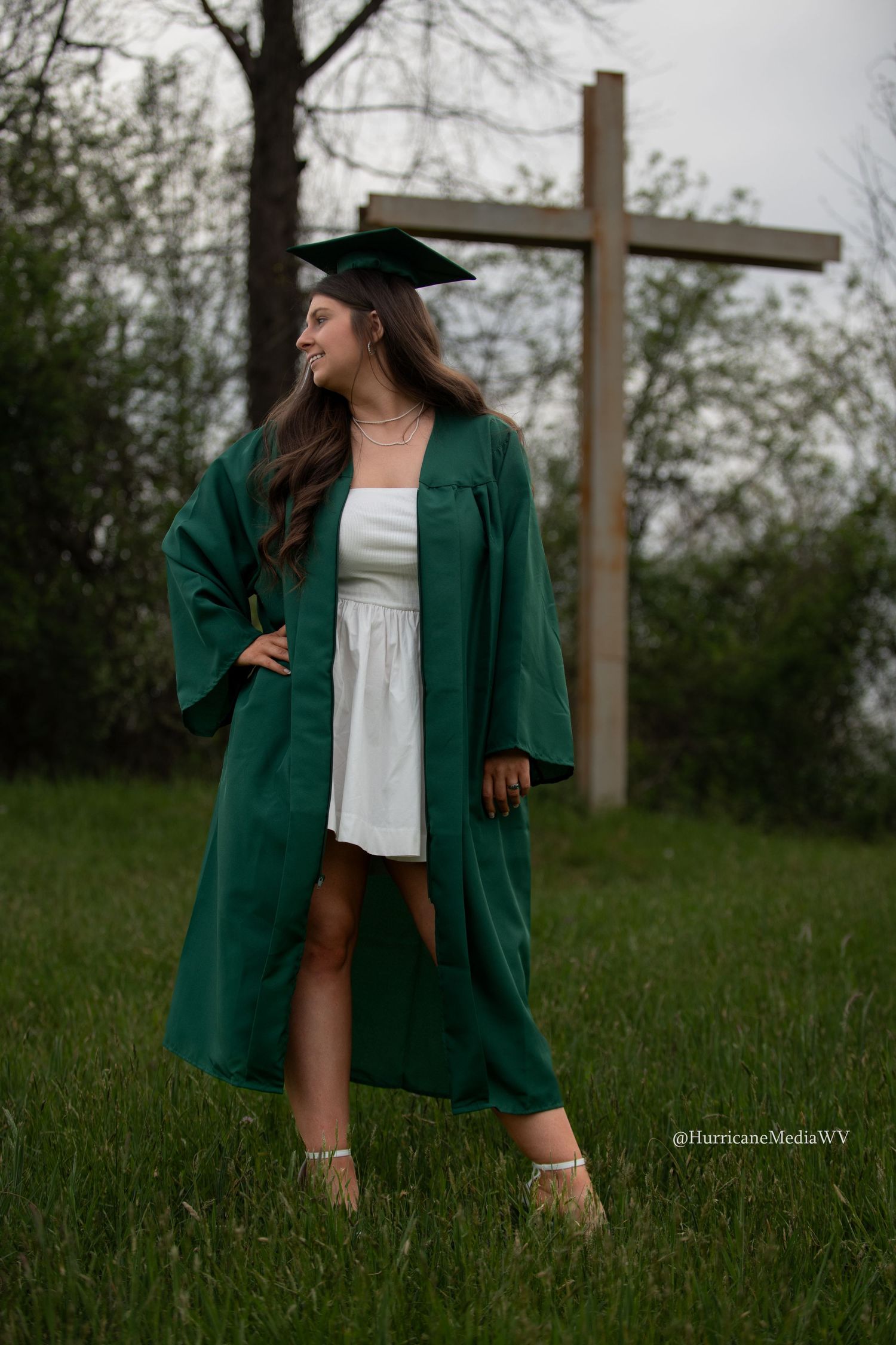 Graduate in emerald green gown and cap poses playfully in a grassy field.