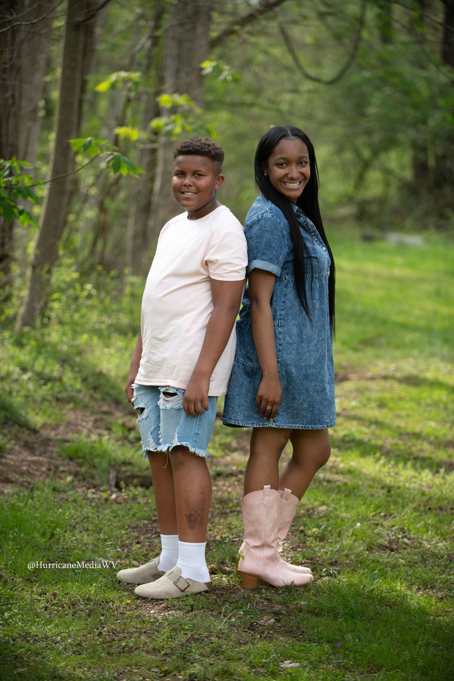Two pairs of legs in casual wear and sneakers standing on grass in a wooded area.