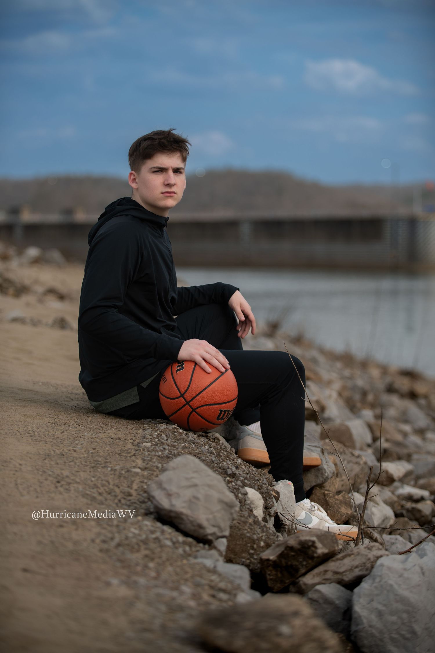 Athlete in black hoodie sits on rocky shore holding basketball against scenic waterfront backdrop.