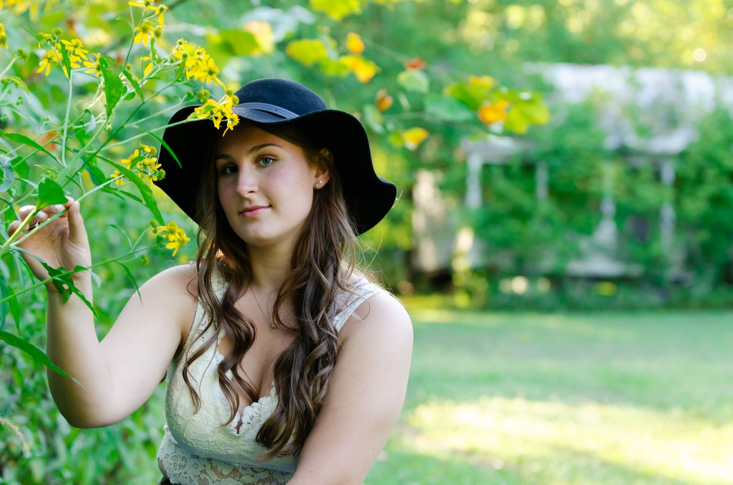 A woman in a black sun hat poses near yellow flowers in a garden setting.