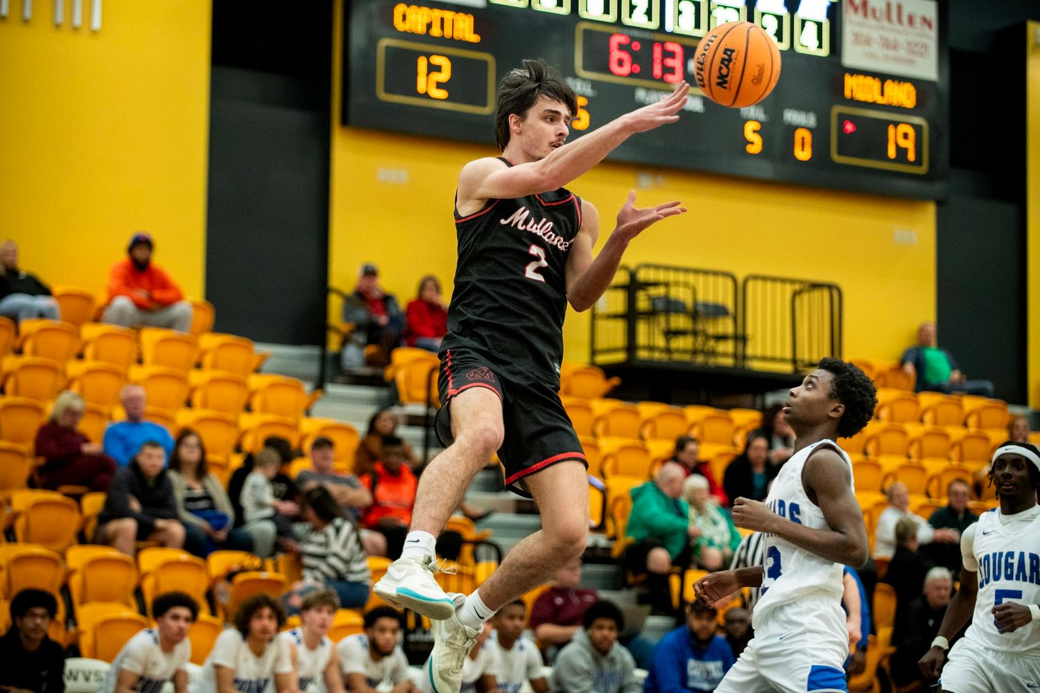 A basketball player leaps high during an indoor game with yellow bleachers in the background.