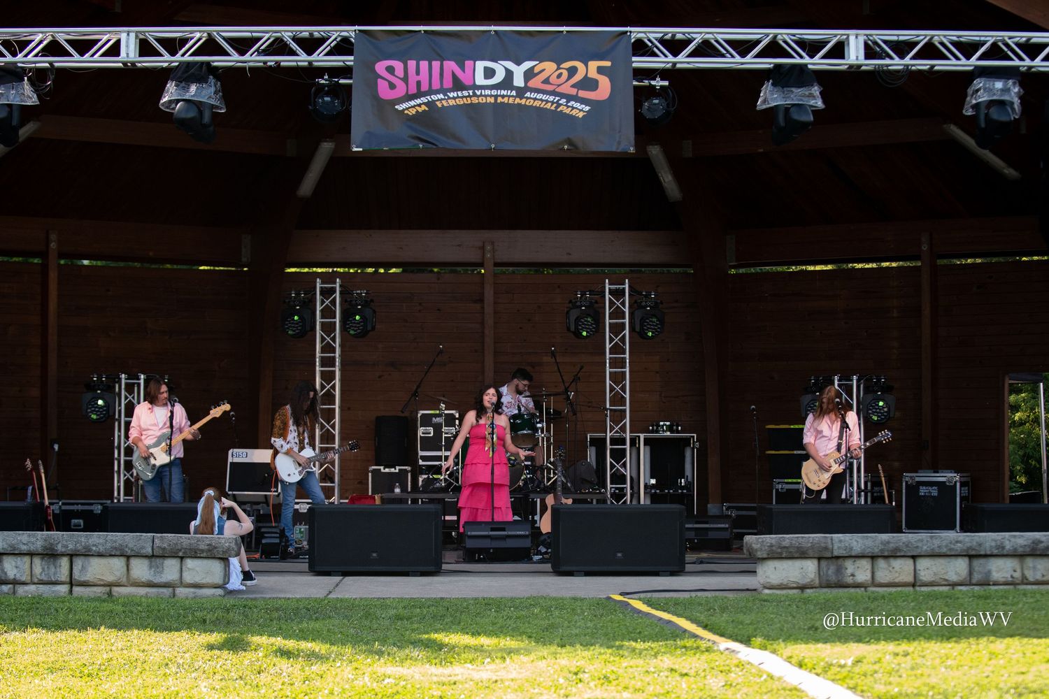 A band performs on an outdoor stage with a 'Shindy' banner overhead during a summer festival performance.