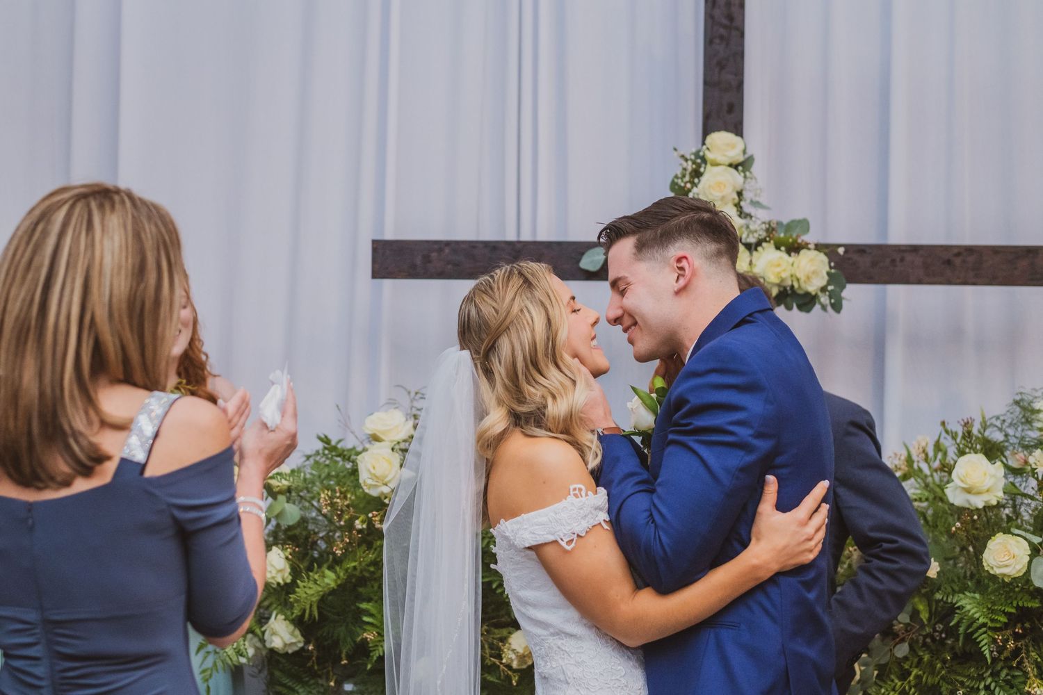 A romantic wedding sequence showing couple embracing and kissing in front of white roses and gray wall backdrop.