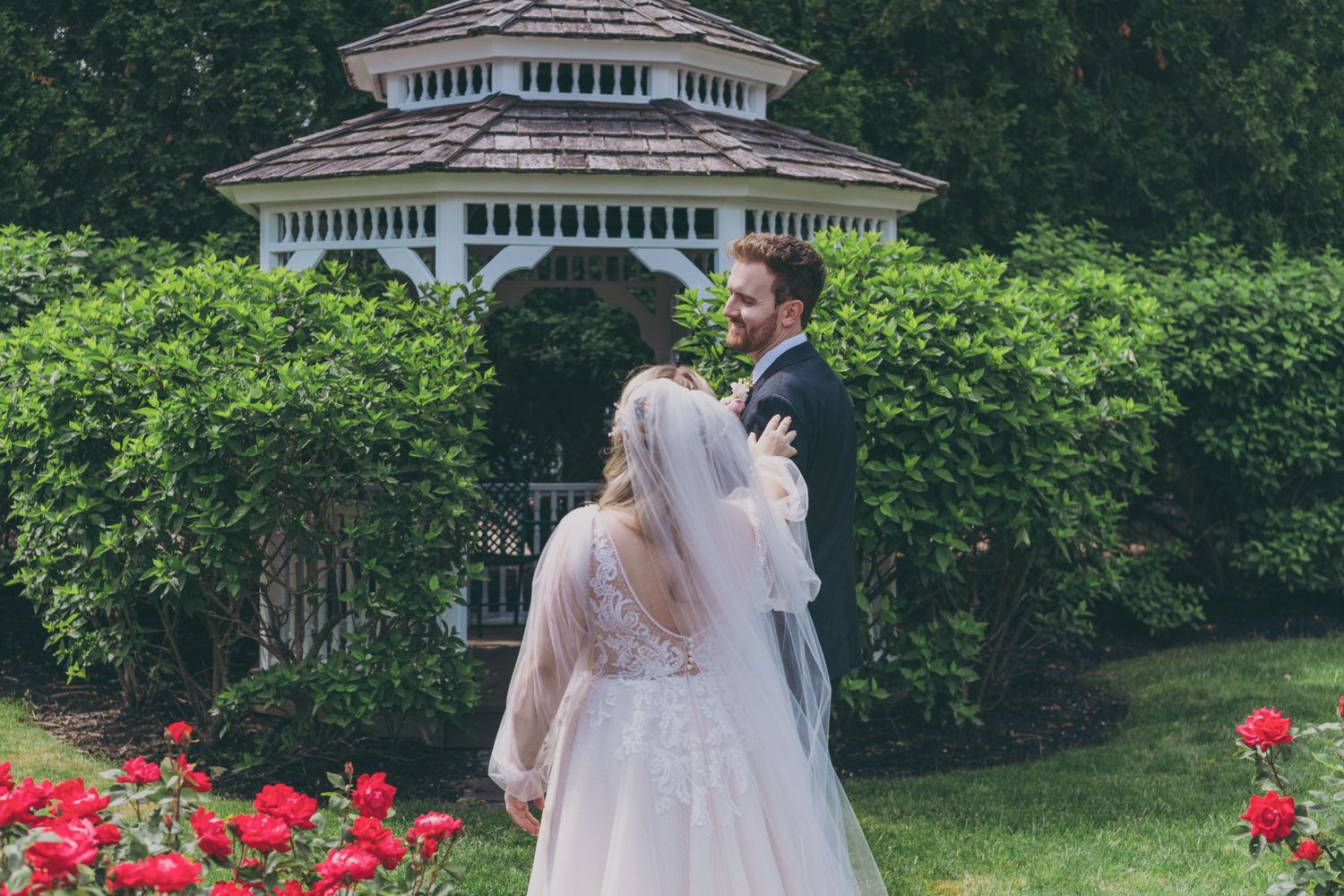 A couple in wedding attire embraces near white gazebo surrounded by lush garden and red rose bushes.