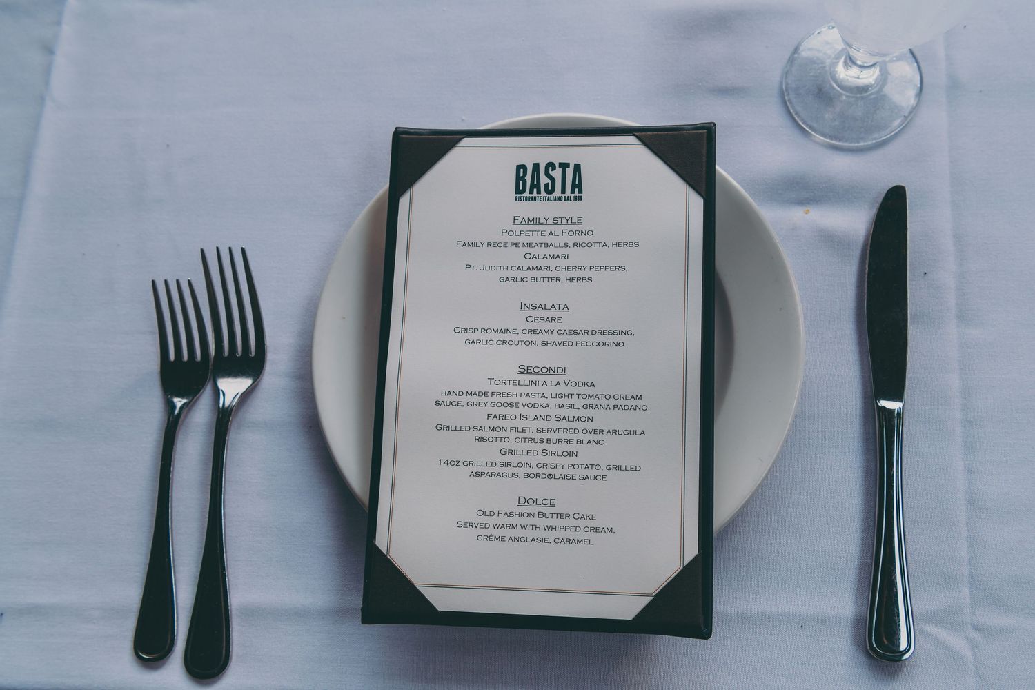 An elegant restaurant menu for 'Basta' sits on a white plate with black cutlery on a white tablecloth.