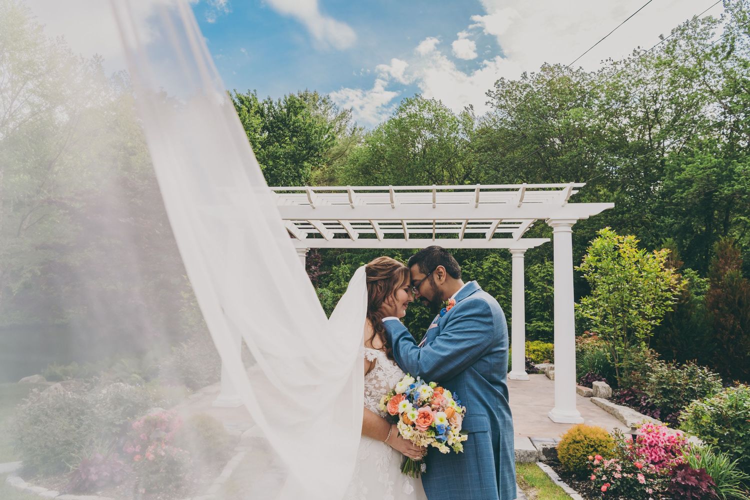A romantic moment shared between a couple under a white pergola surrounded by flowers on a sunny day.