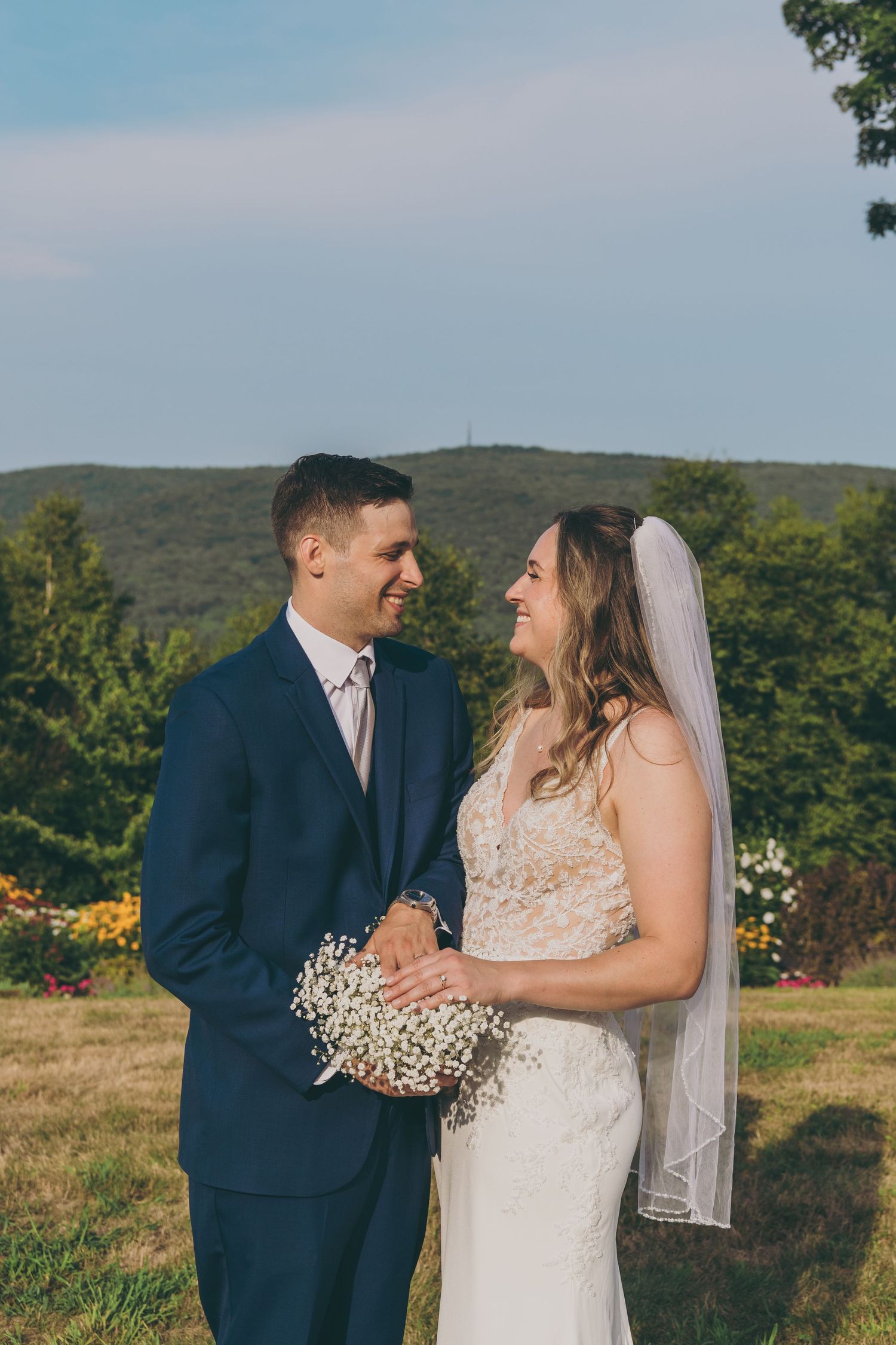 Couple in wedding attire share joyful moment against mountain backdrop with garden flowers.