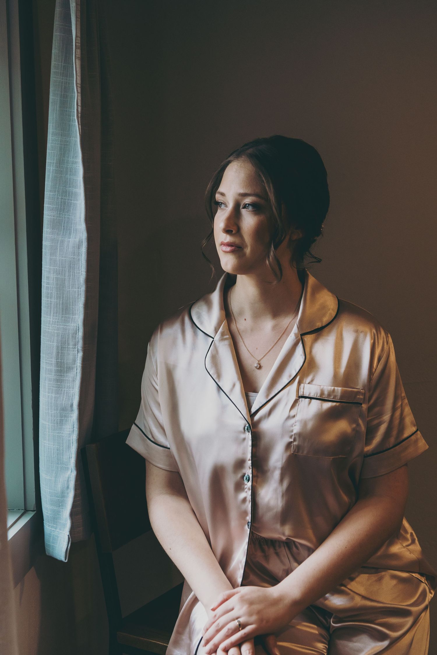 Person wearing elegant white satin pajamas sits by window in moody natural lighting.