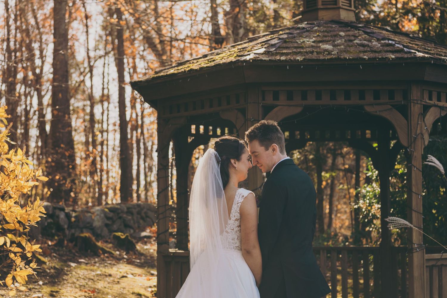 A couple in wedding attire poses under an autumn gazebo as golden leaves fall in a scenic outdoor setting.