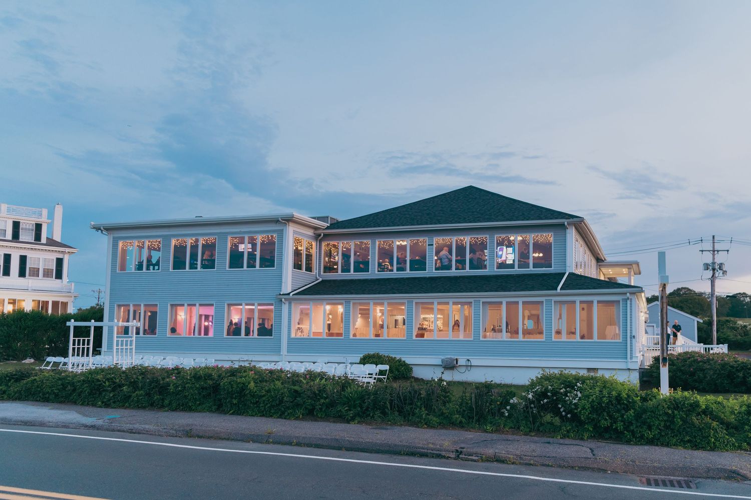 A light blue coastal restaurant with large windows glows at dusk along a seaside road.