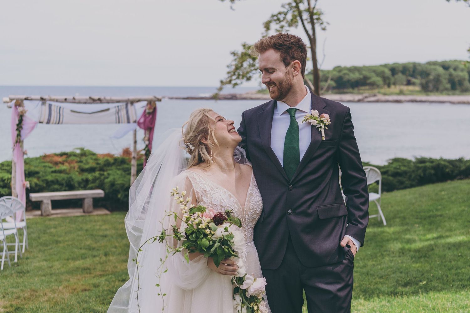 A joyful wedding couple sharing a candid moment together on a grassy lawn overlooking the water.