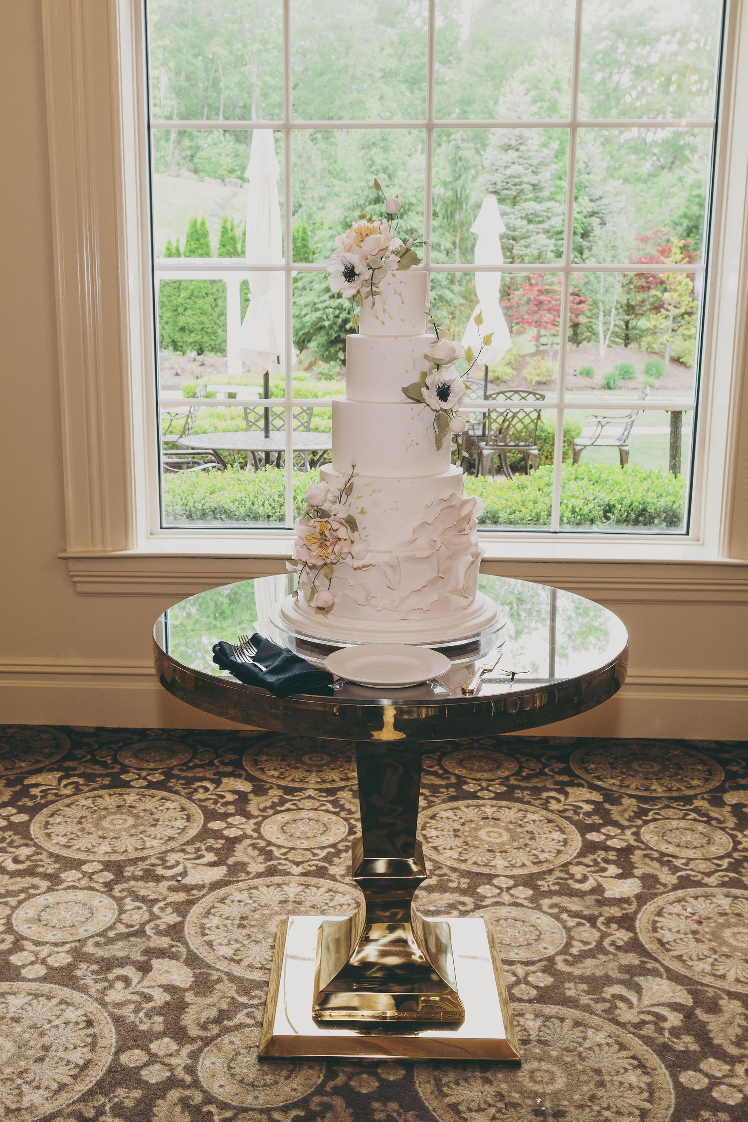 Elegant white wedding cake with floral decorations displayed on ornate black and gold pedestal table by garden window.