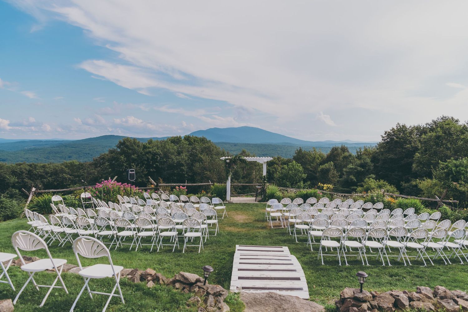 White chairs arranged for an outdoor mountain view wedding ceremony with a wooden aisle on a scenic hilltop location.