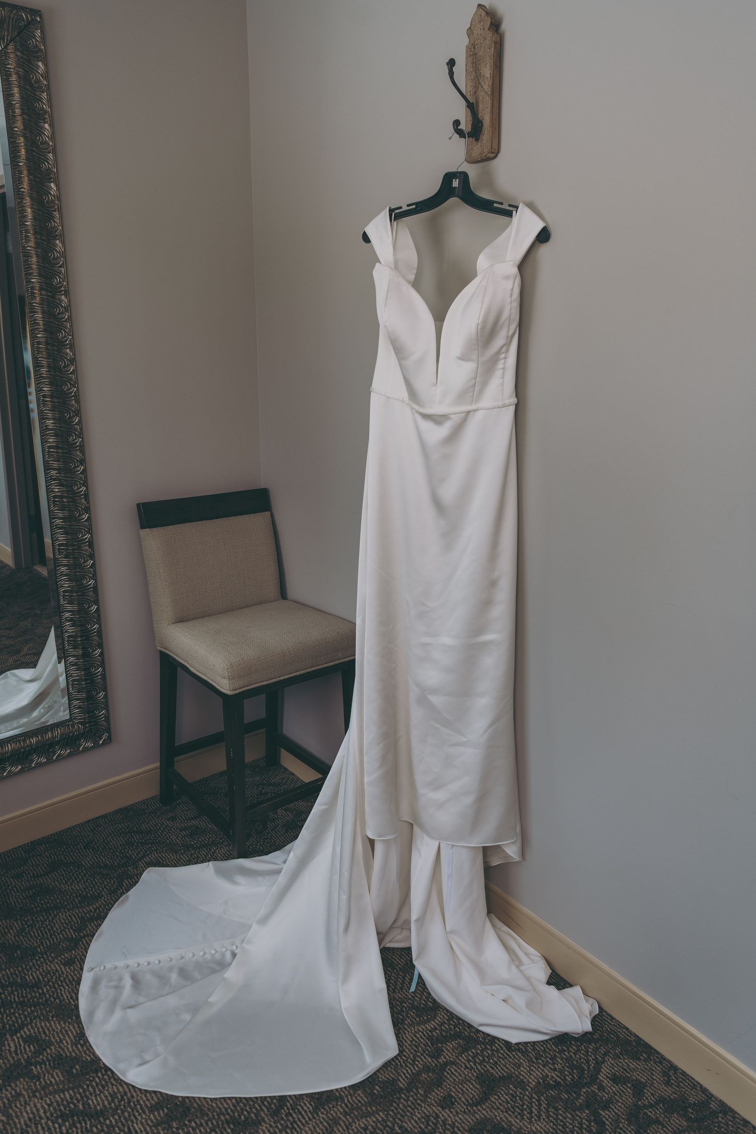 An elegant white wedding dress with a plunging neckline and long train hanging on a hanger in a corner room.