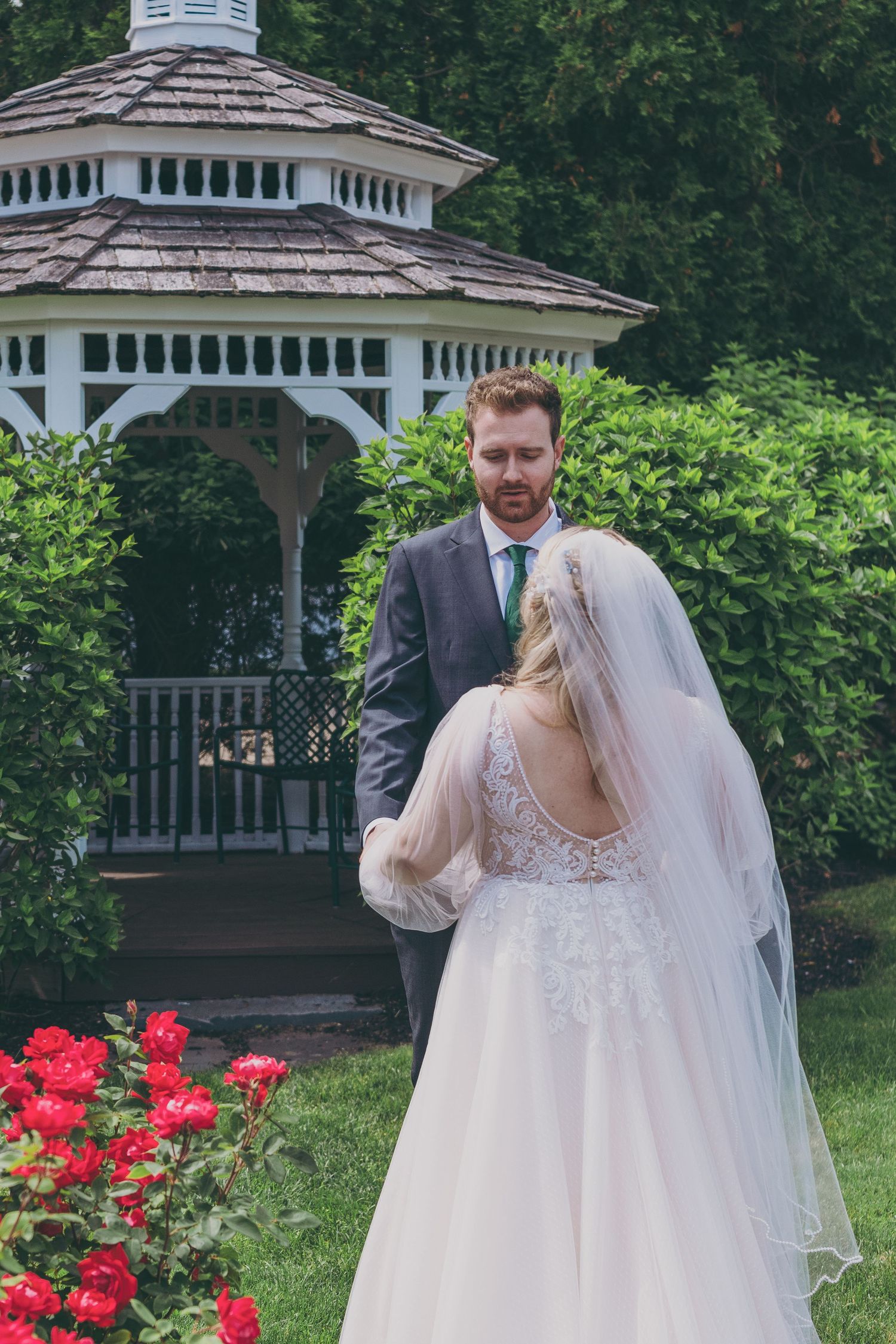 Bride and groom share a tender moment by a garden gazebo with red roses in bloom on a verdant summer day.