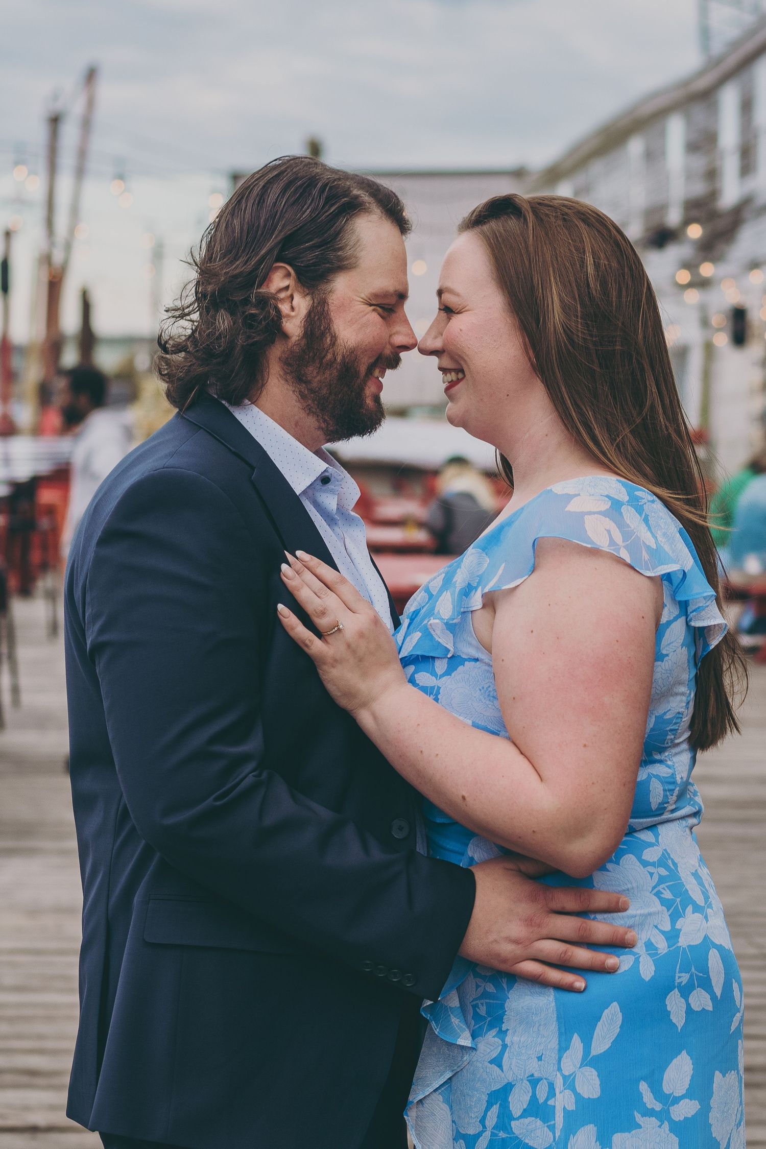 A romantic couple embracing on a boardwalk, her in blue floral dress and him in navy suit.