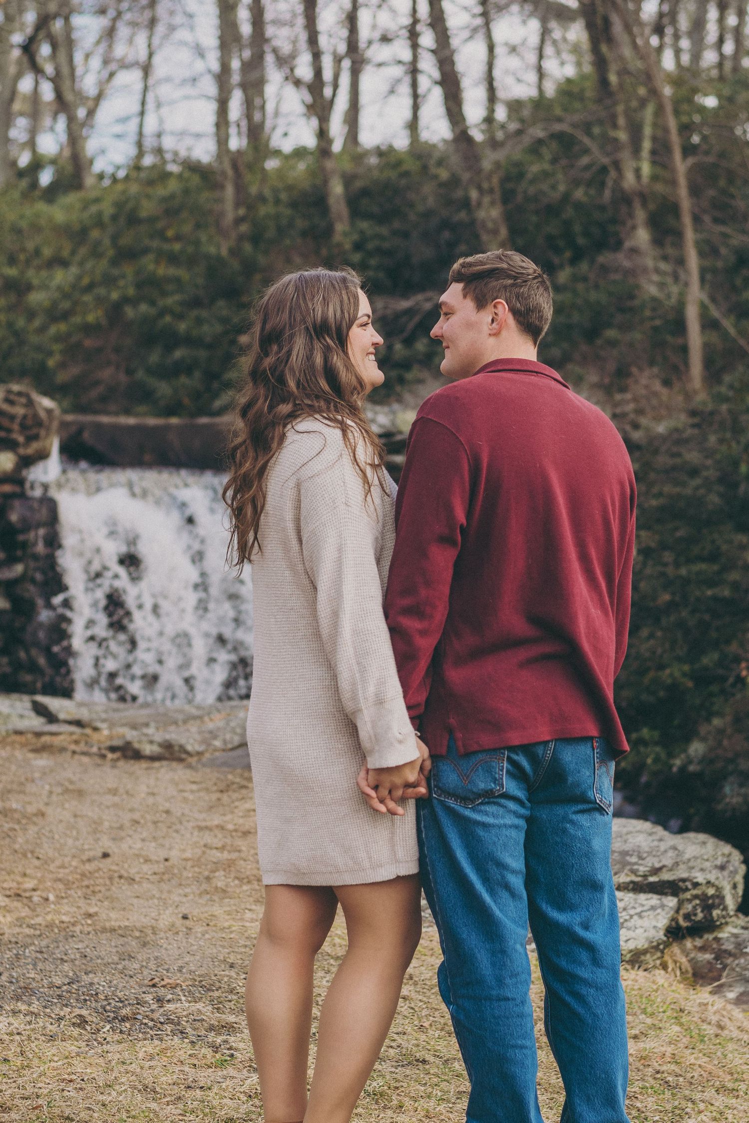 Couple holding hands near a waterfall, dressed in casual winter attire against a wooded backdrop.