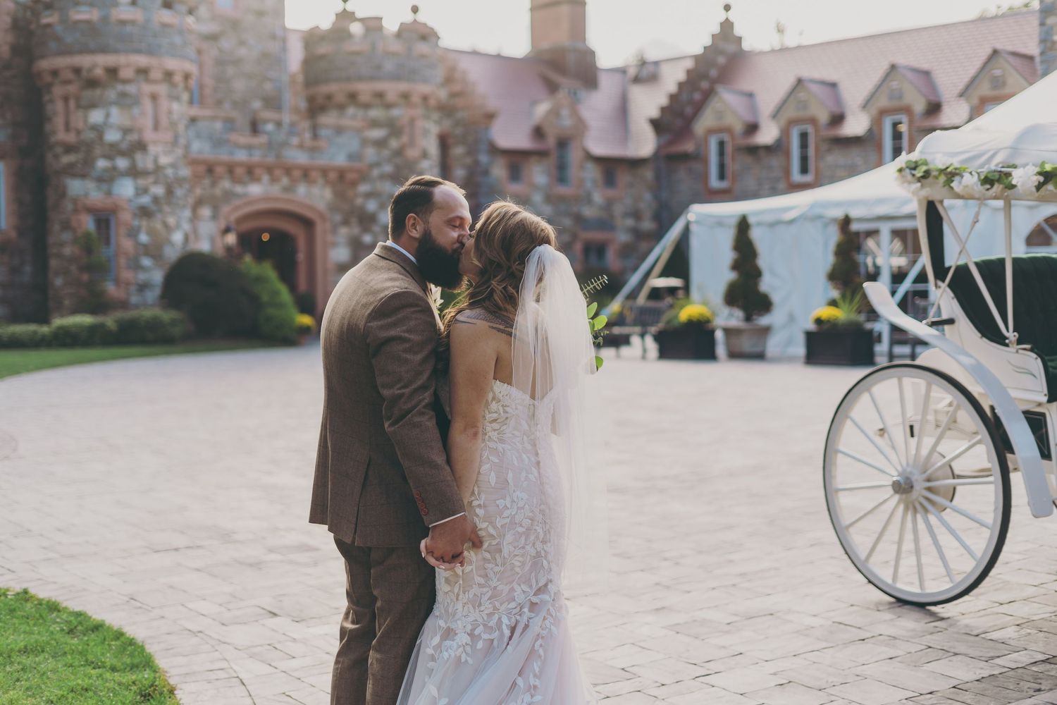 A romantic wedding kiss takes place in front of a stone castle with a vintage carriage wheel visible in the background.