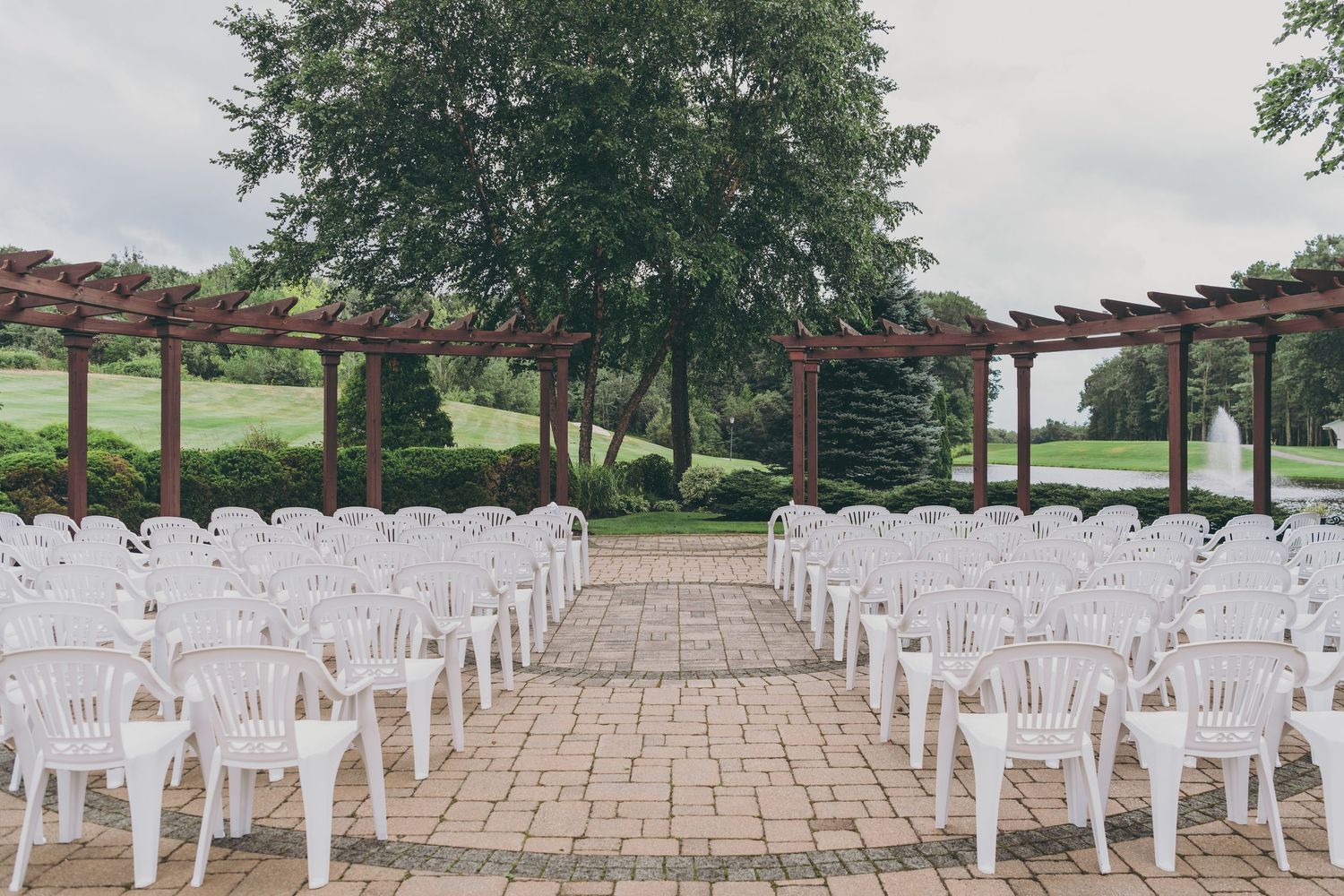 White chairs line a stone pathway leading to wooden pergolas in an outdoor garden wedding ceremony setup.