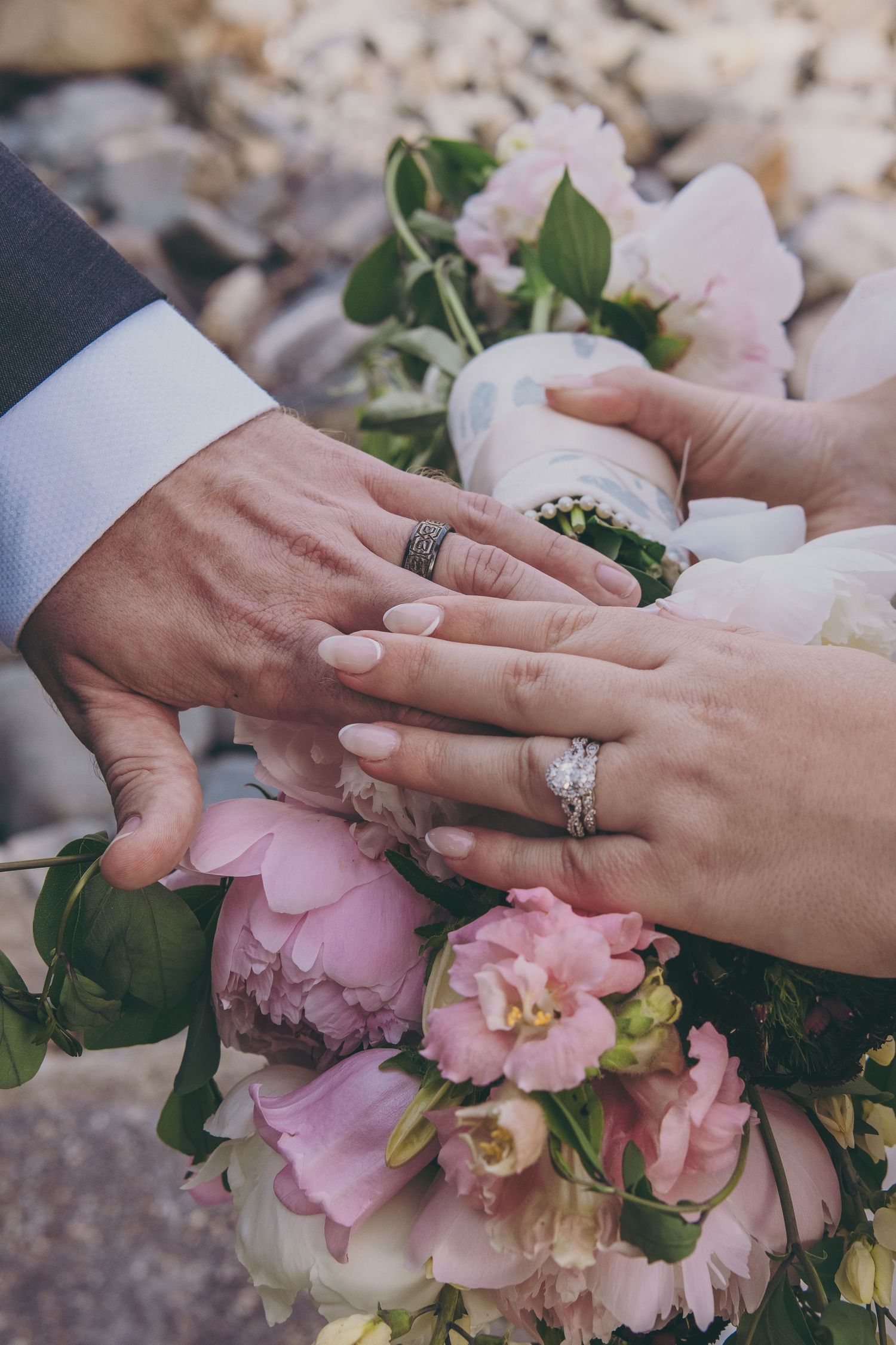 Newlywed hands displaying wedding rings over a bouquet of soft pink peonies and roses.