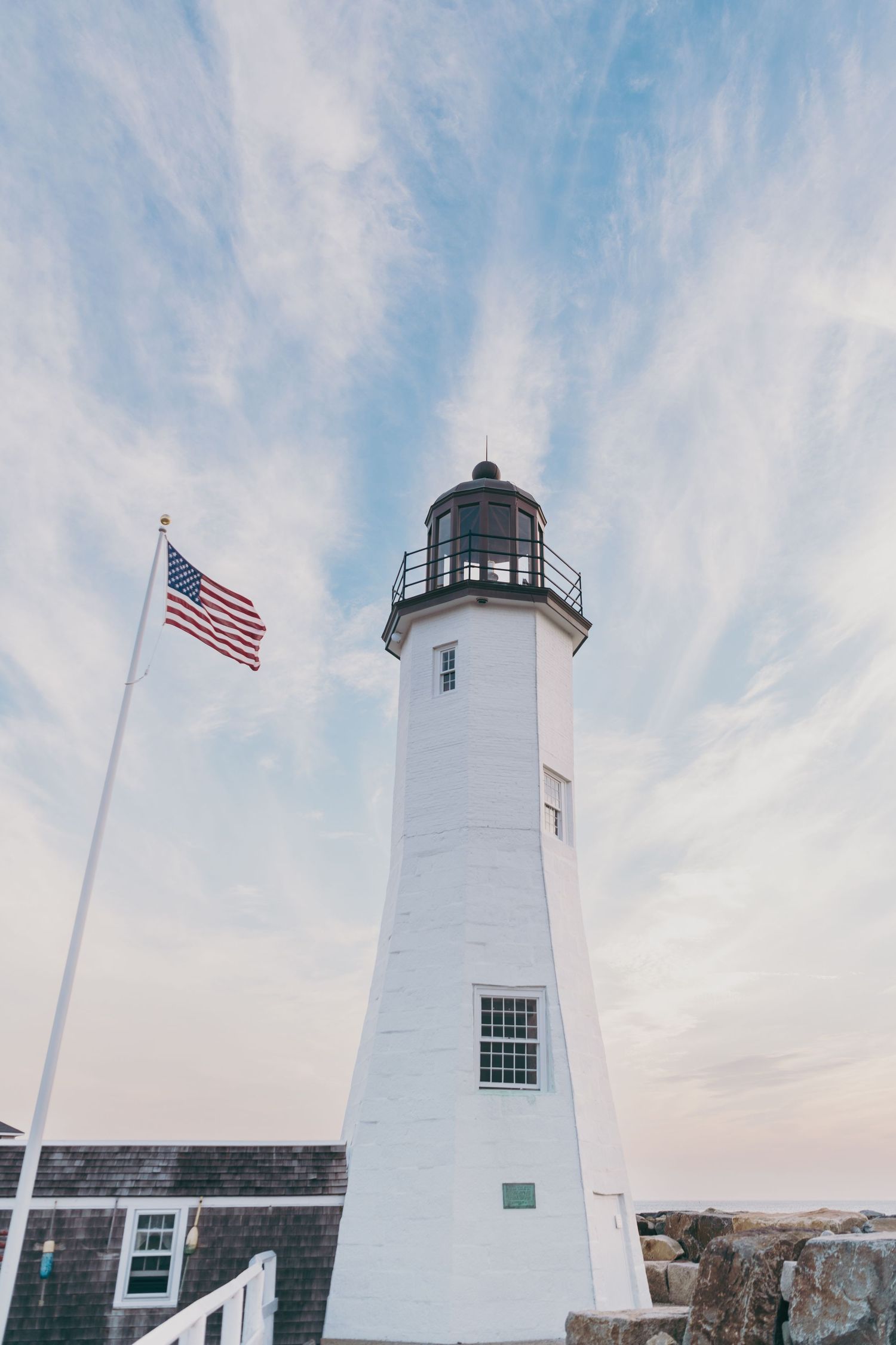 Tall white lighthouse tower with American flag flying against cloudy blue sky at Scituate Light in Massachusetts.