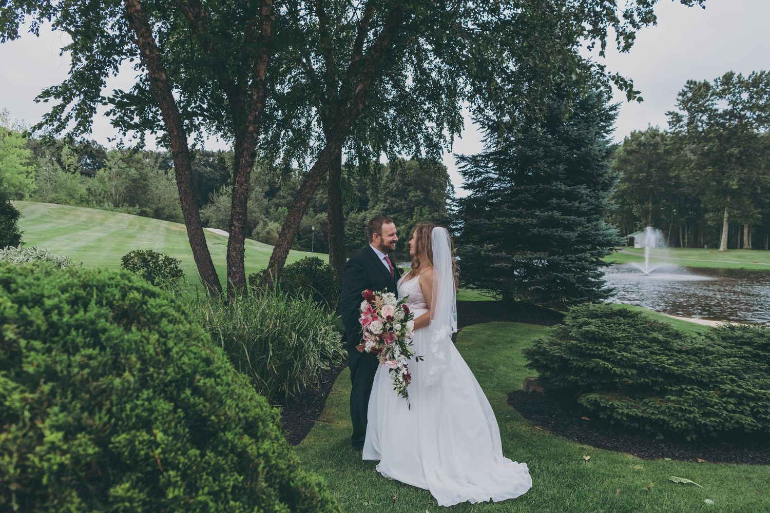 A couple stands together on a lush golf course with a water feature and evergreen trees in the background.