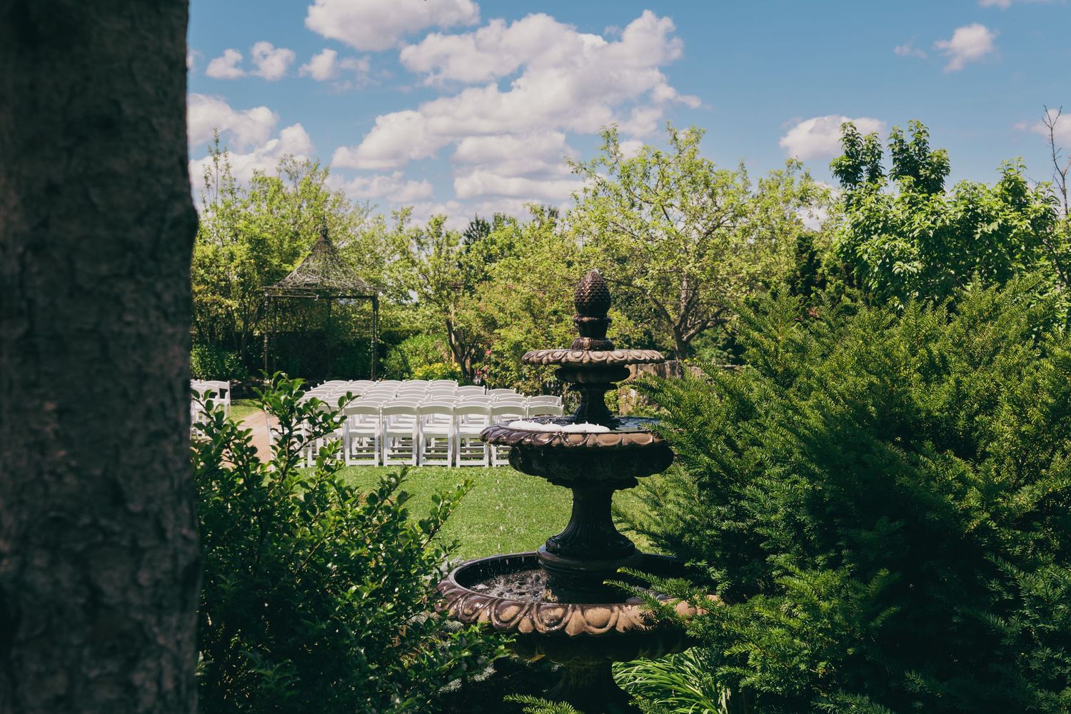A decorative stone fountain sits in a lush garden surrounded by green trees and shrubs under a blue cloudy sky.