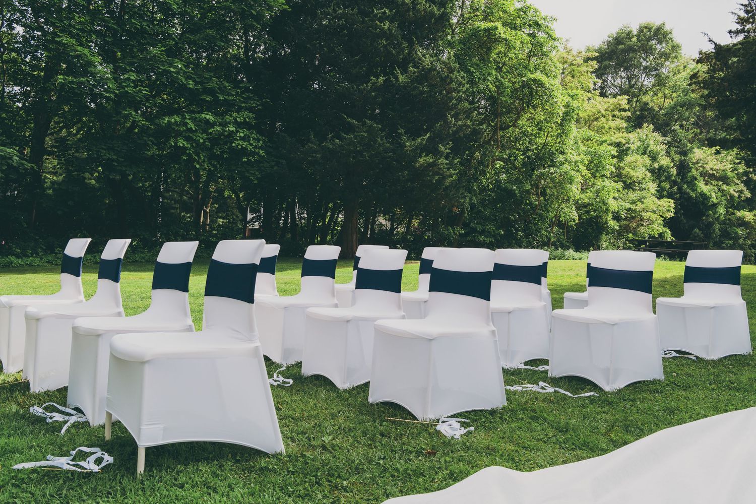 White tablecloths drape over outdoor event tables arranged on a lush green lawn beneath tall trees.