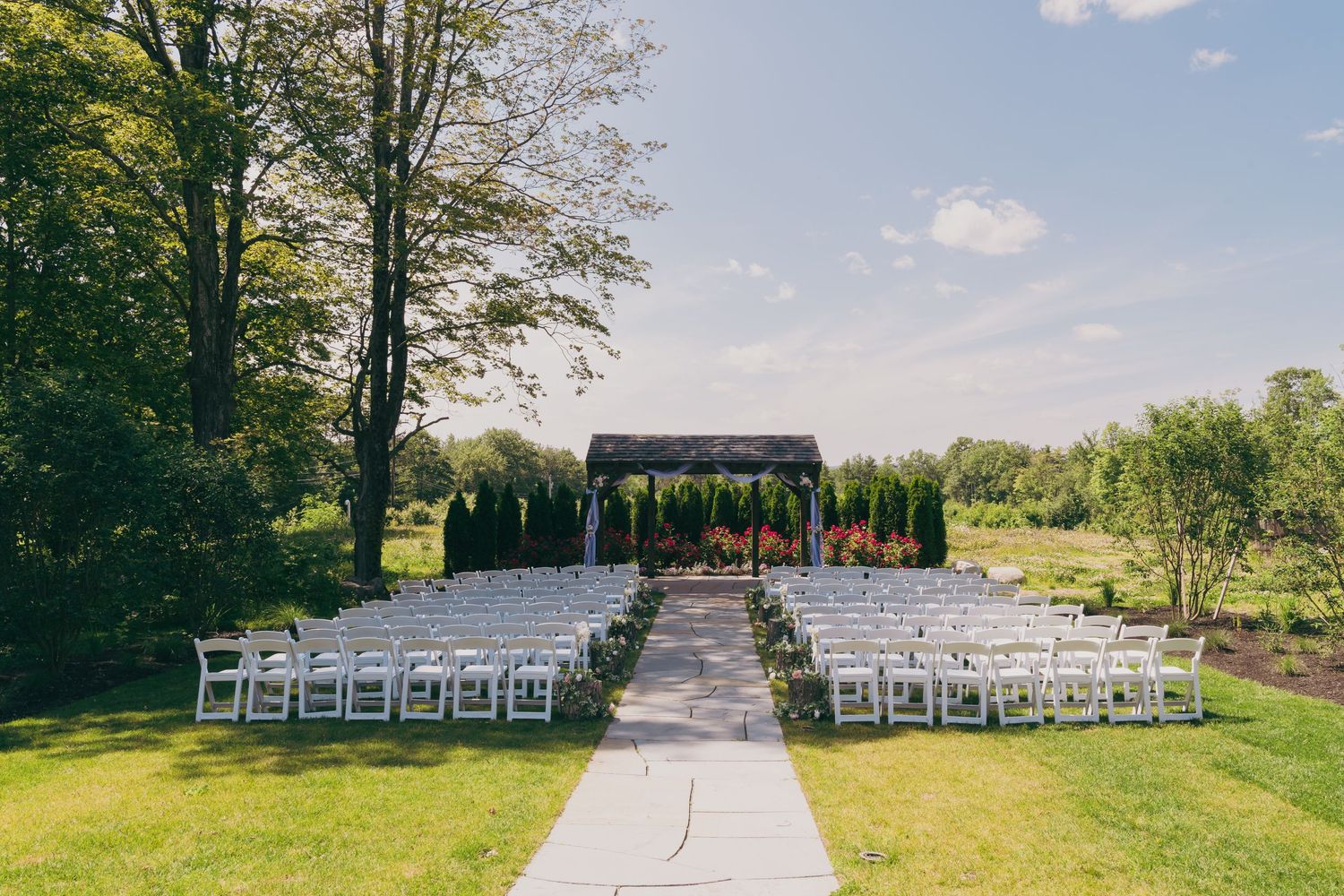 Outdoor wedding venue setup with white chairs arranged in rows facing a wooden arch against a natural backdrop.