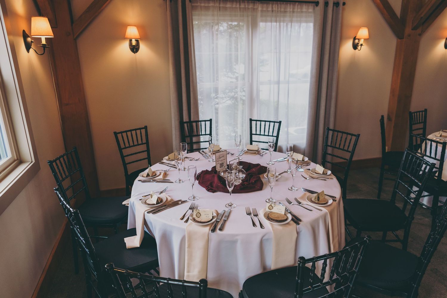 A round dining table with white tablecloth and burgundy accents, set with formal place settings and surrounded by dark chairs.