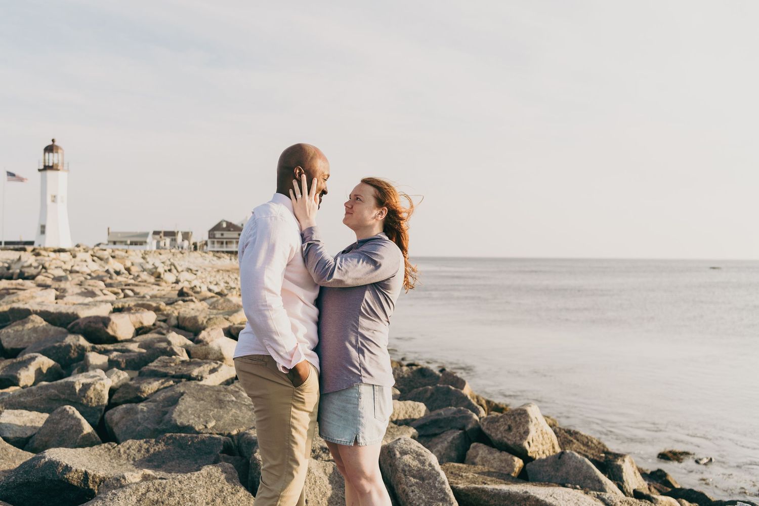 A romantic couple embraces on rocky shore with lighthouse in background along misty coastline.