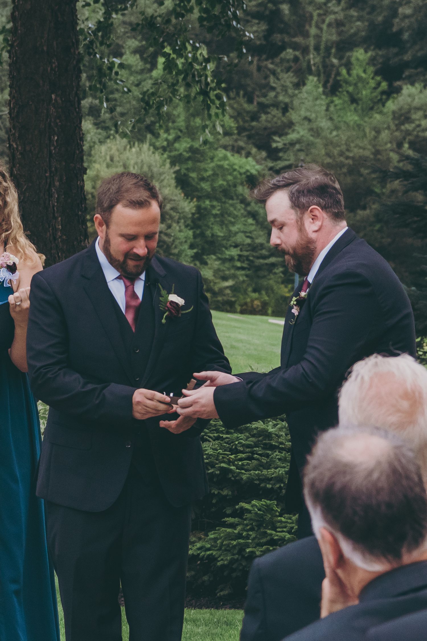 Two men in formal suits exchange rings during an outdoor wedding ceremony.