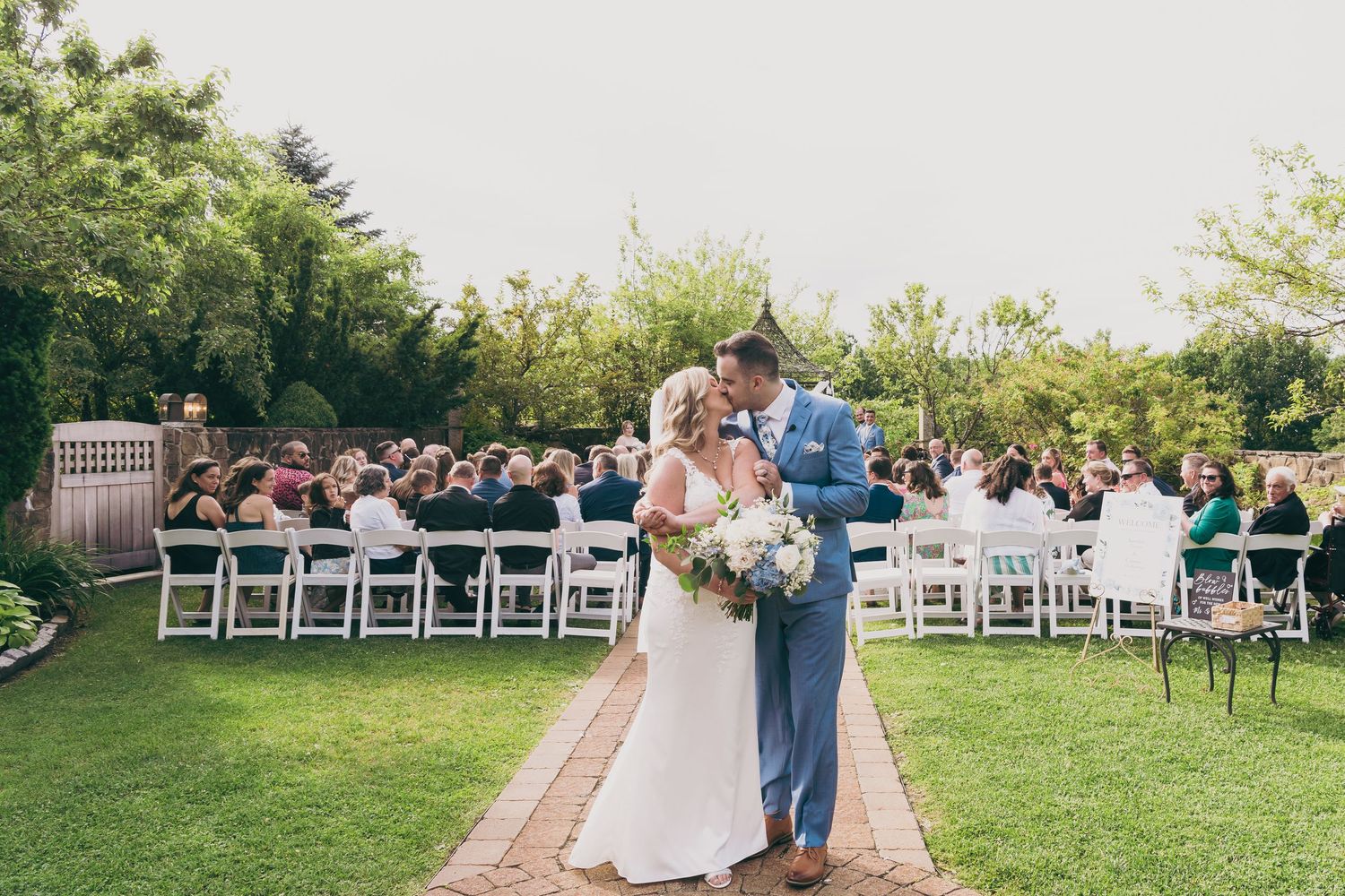 Outdoor garden wedding ceremony with white chairs and couple standing on stone pathway during daytime celebration at the Granite Rose.