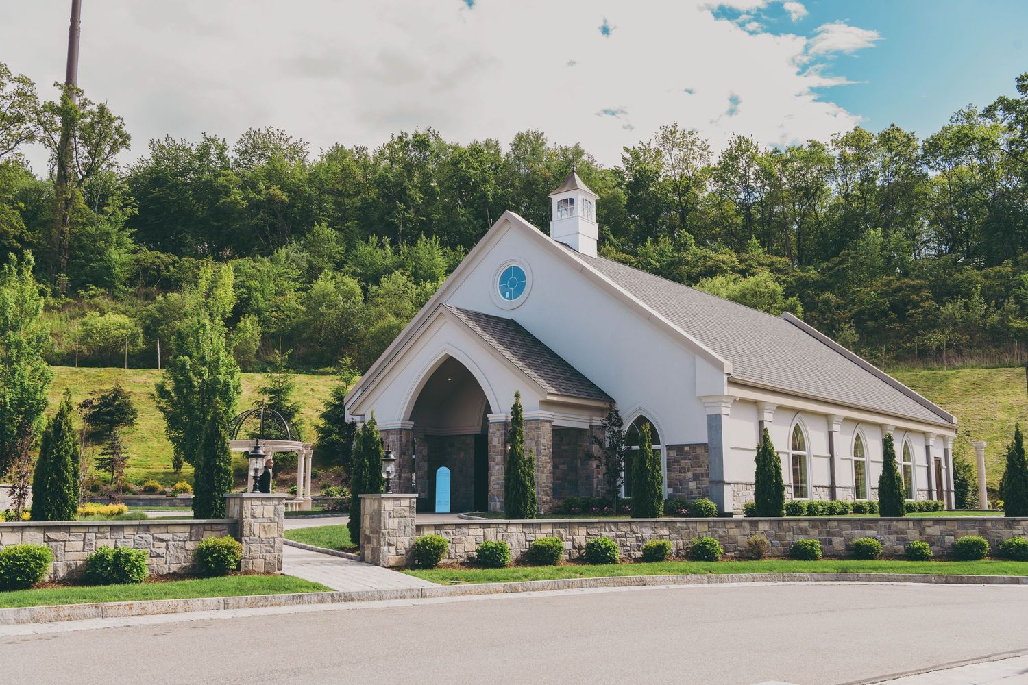 A charming white country chapel with arched entrance and blue door sits nestled against rolling green hills on a sunny day.